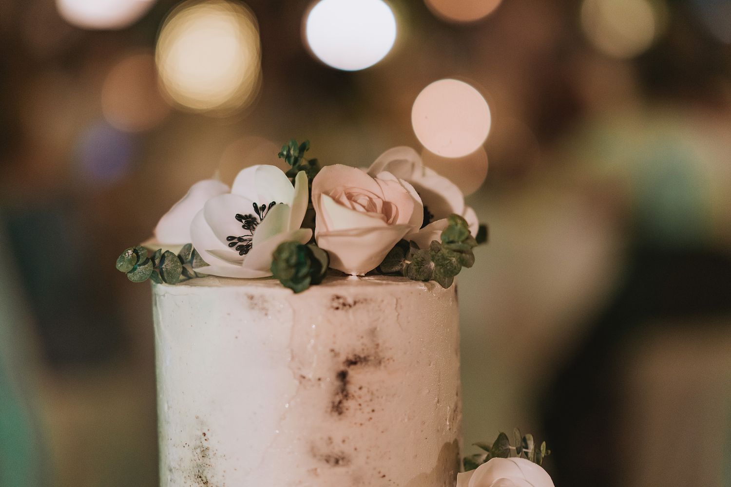 Close-up of an elegant white wedding cake decorated with blush pink roses and greenery on top with bokeh lights in background.