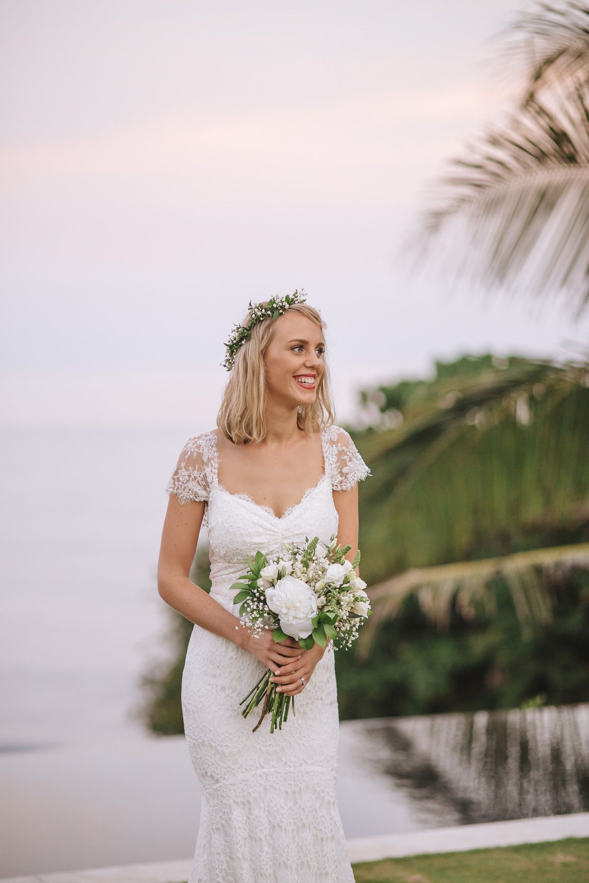 A radiant bride in a lace dress and flower crown holds a bouquet against a tropical sunset backdrop.