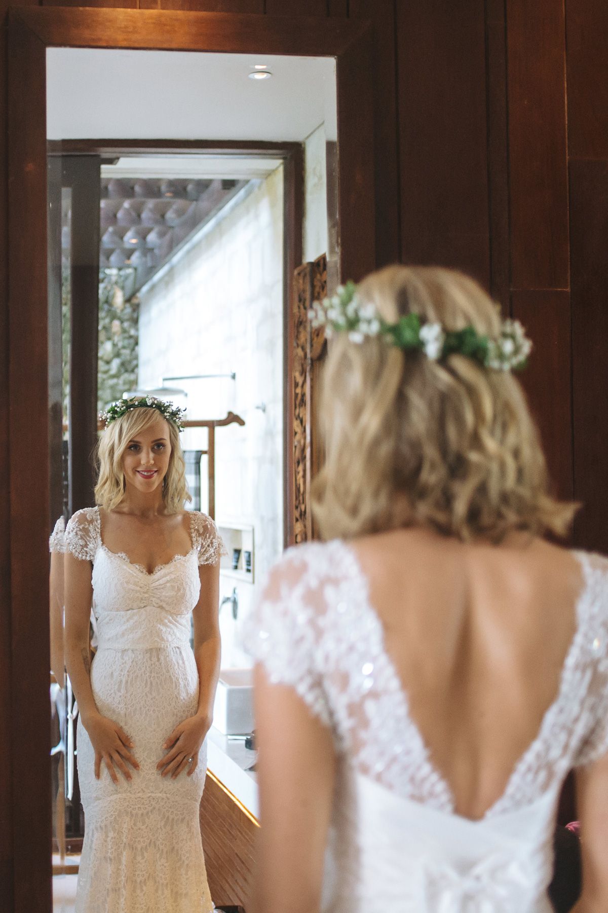 Bride smiling in white lace wedding dress with floral crown, seen in multiple mirror reflections.