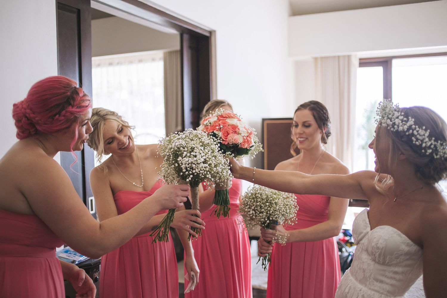 Wedding party in coral pink dresses admiring bridal bouquets filled with baby's breath flowers indoors.
