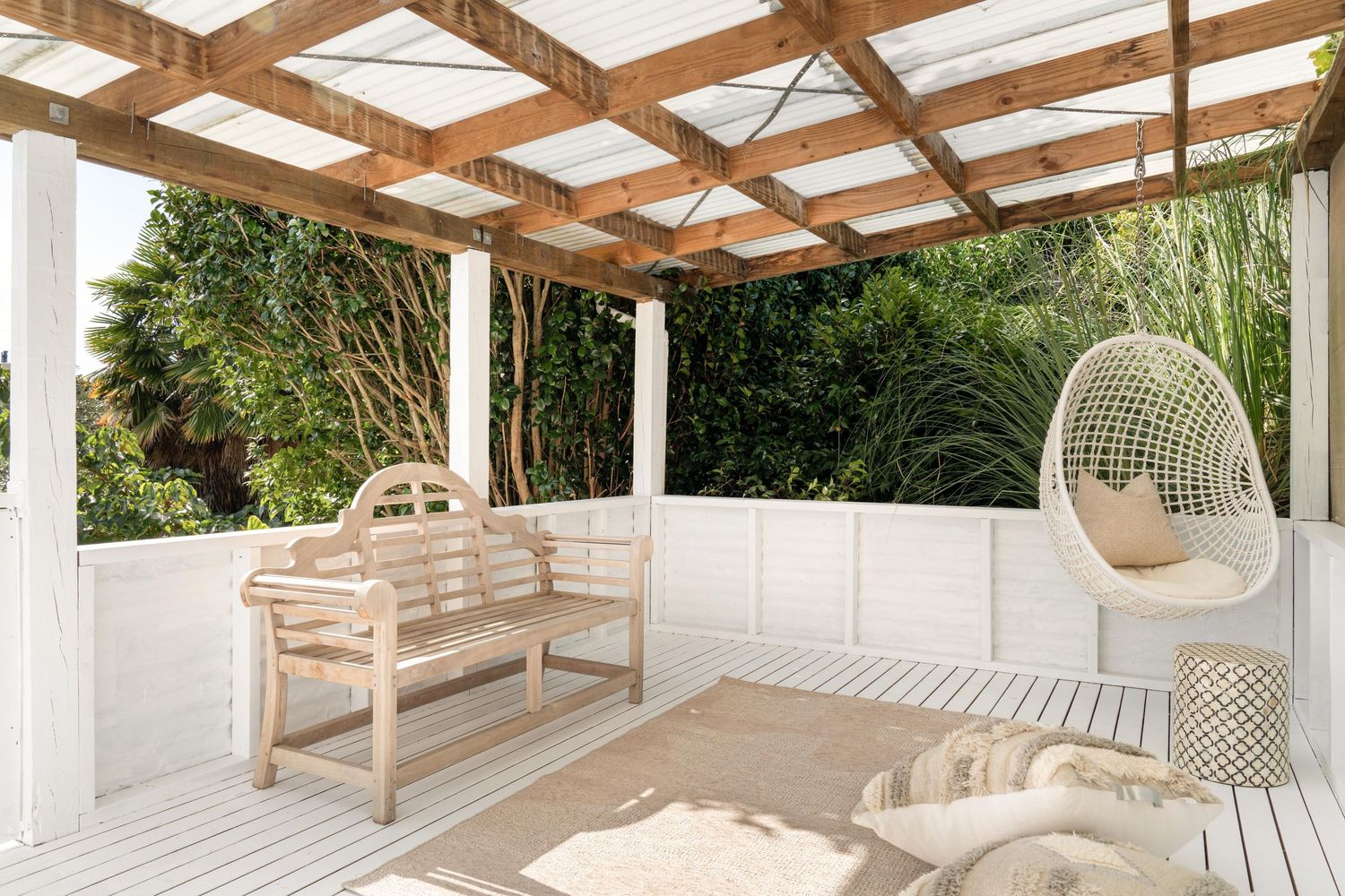 Bright outdoor patio with wooden pergola, white benches and a hanging egg chair.