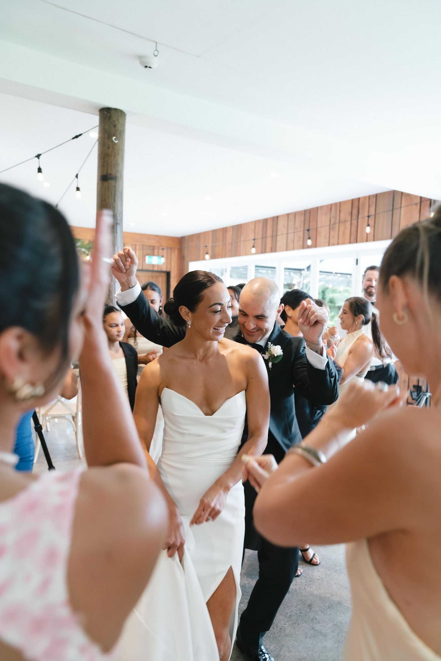 A lively wedding reception dance party underway in a rustic wood-paneled venue with string lights.