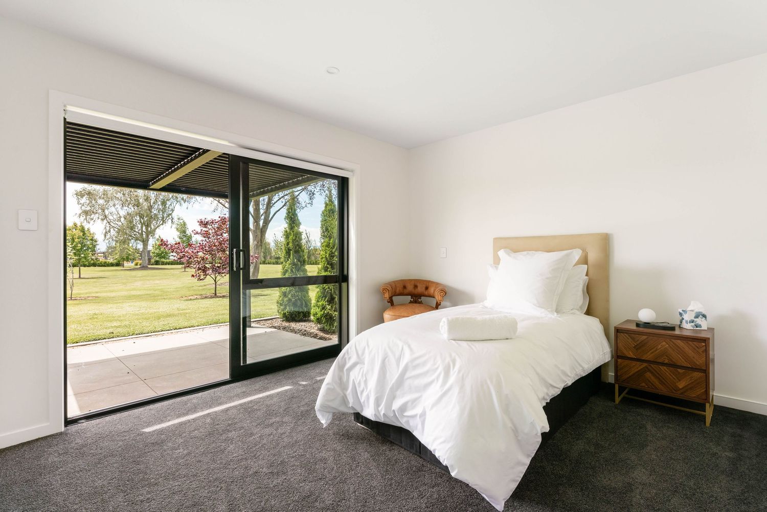 Contemporary bedroom with sliding glass doors, white bedding, and wooden furniture overlooking outdoor patio.