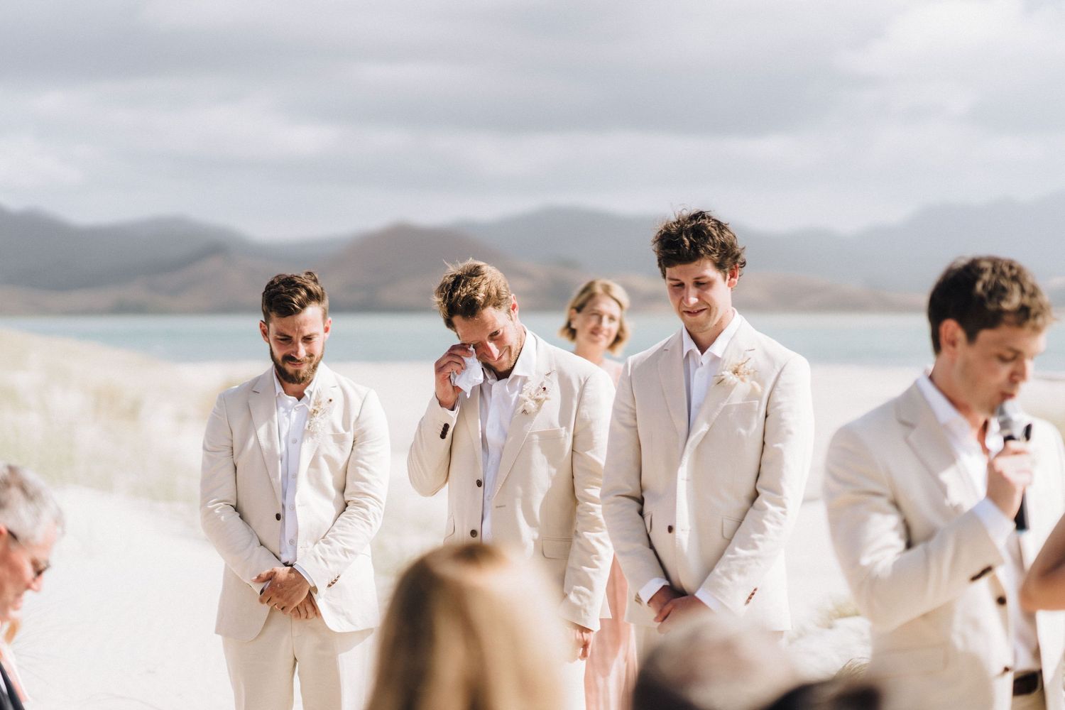 Wedding party wearing white stands together on a sandy beach against cloudy mountain landscape.