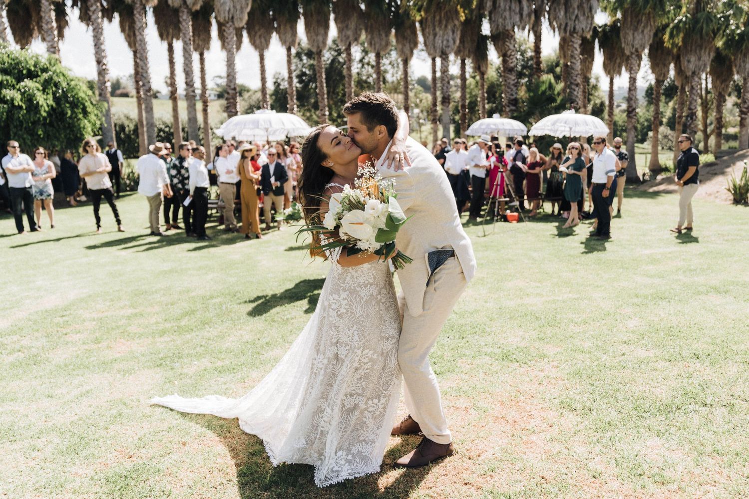 Newlyweds share romantic moment on grassy lawn lined with palm trees while wedding guests look on.