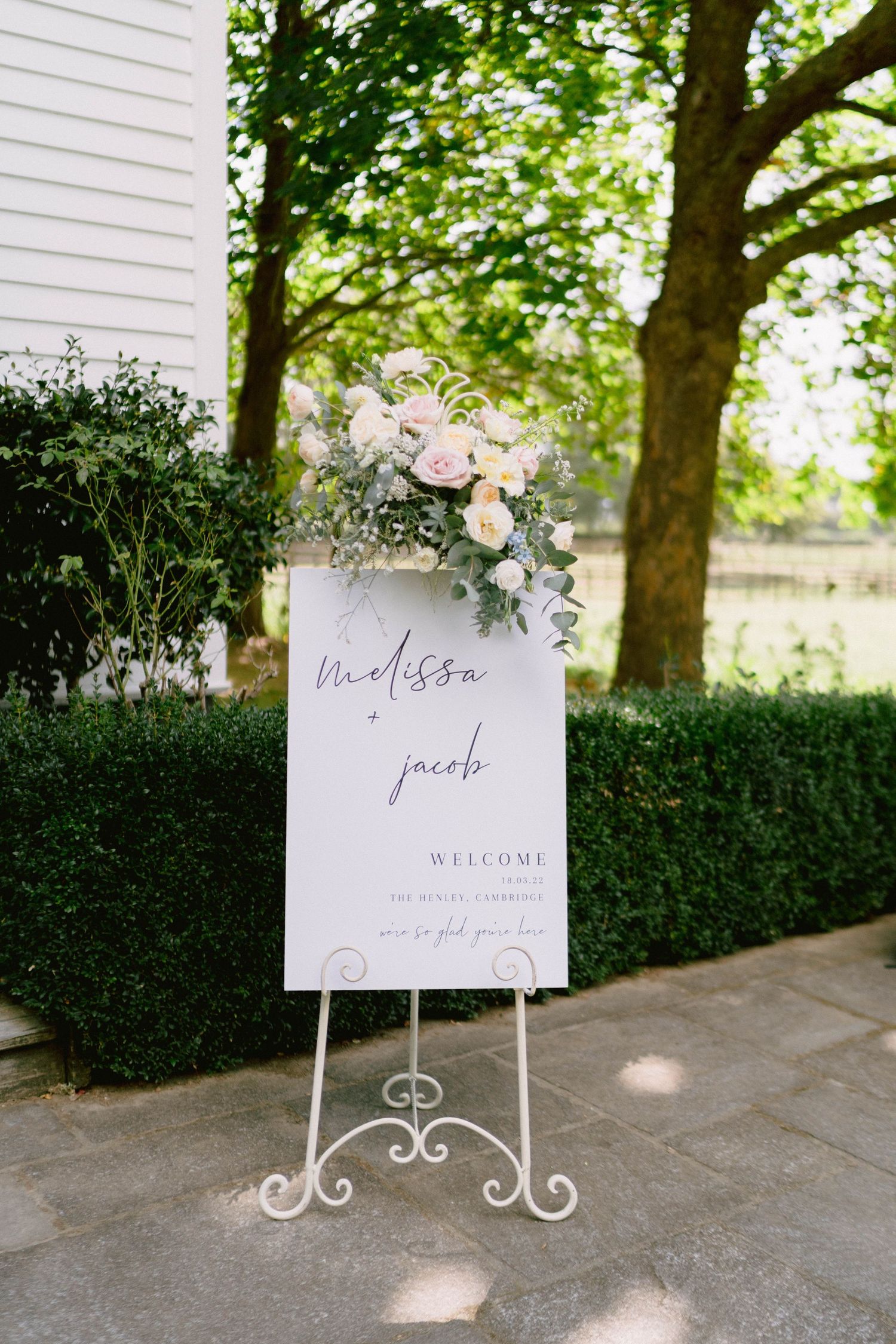 Elegant white wedding welcome signs decorated with blush florals and greenery stand in a garden setting.