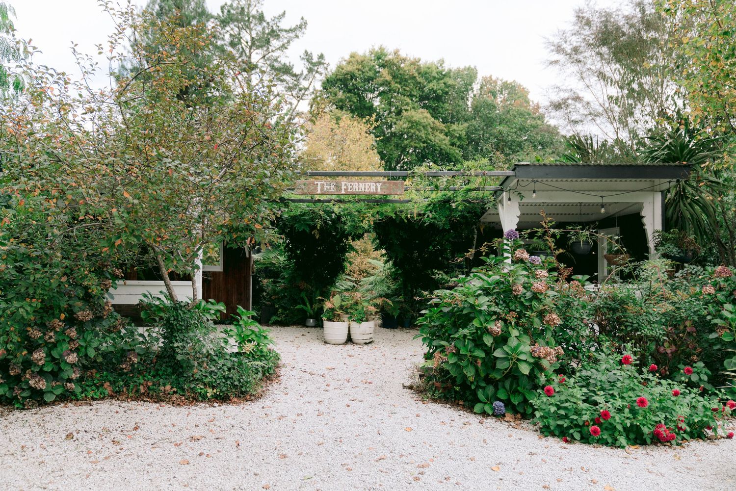 A gravel garden path lined with lush green bushes and trees leads to a covered outdoor structure.