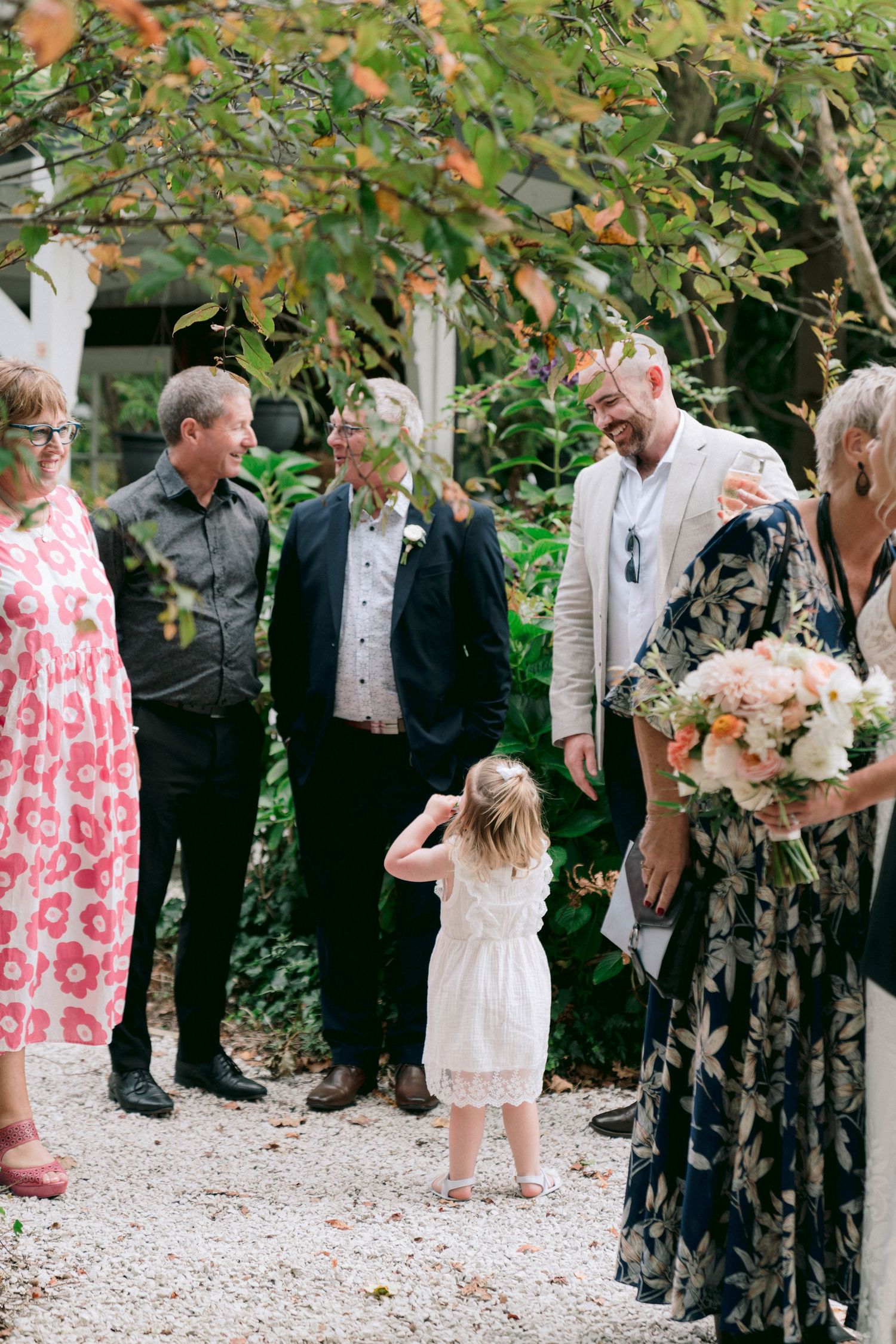 A group gathers in a garden setting while a small child in a white dress stands among them.