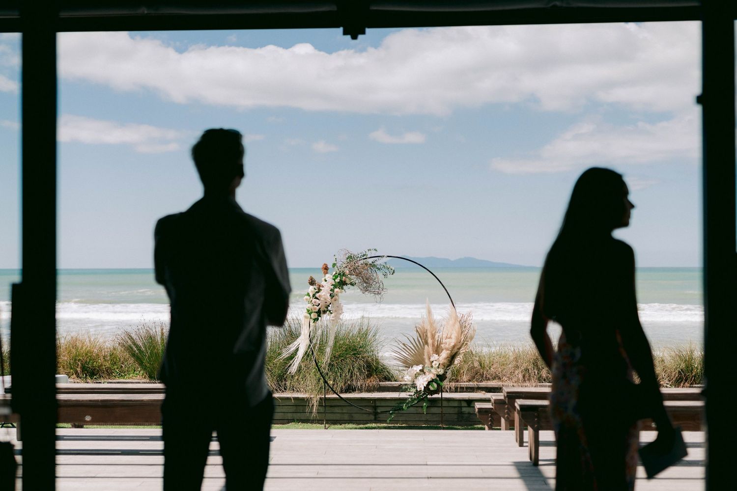 Two silhouettes stand apart against a bright ocean backdrop through large windows.