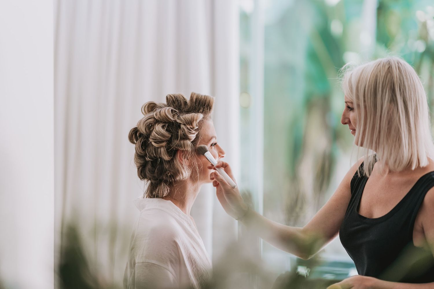 Stylist working on client's hair in elegant salon with natural lighting and white curtains.
