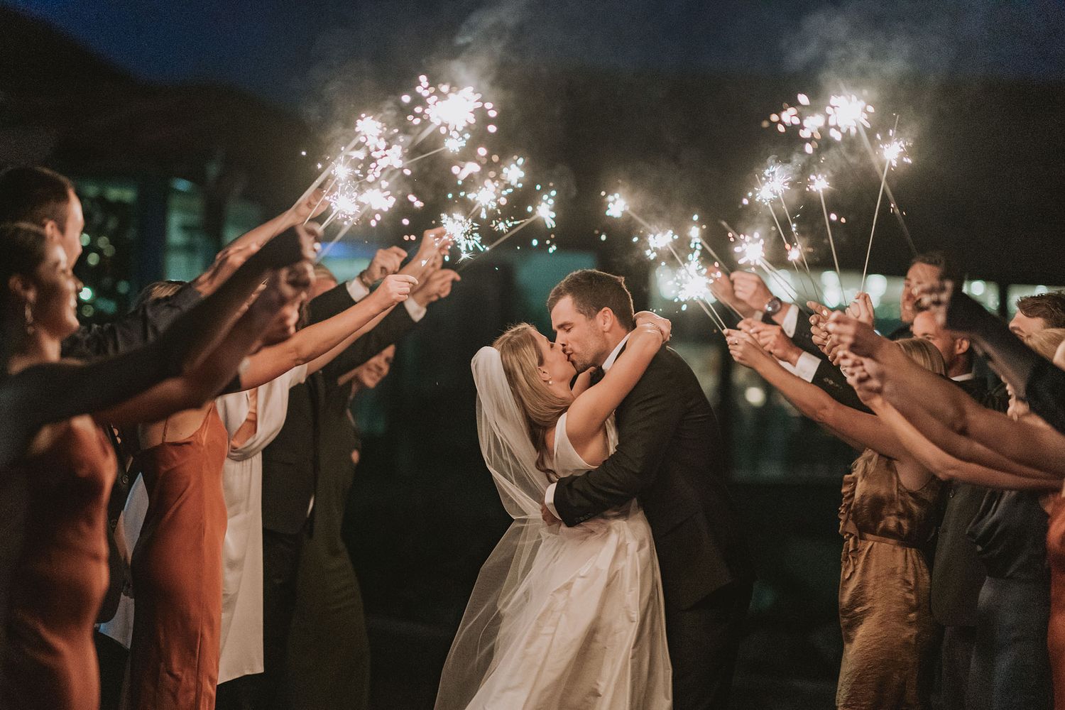 Couple shares romantic kiss under glowing sparklers during nighttime wedding celebration.