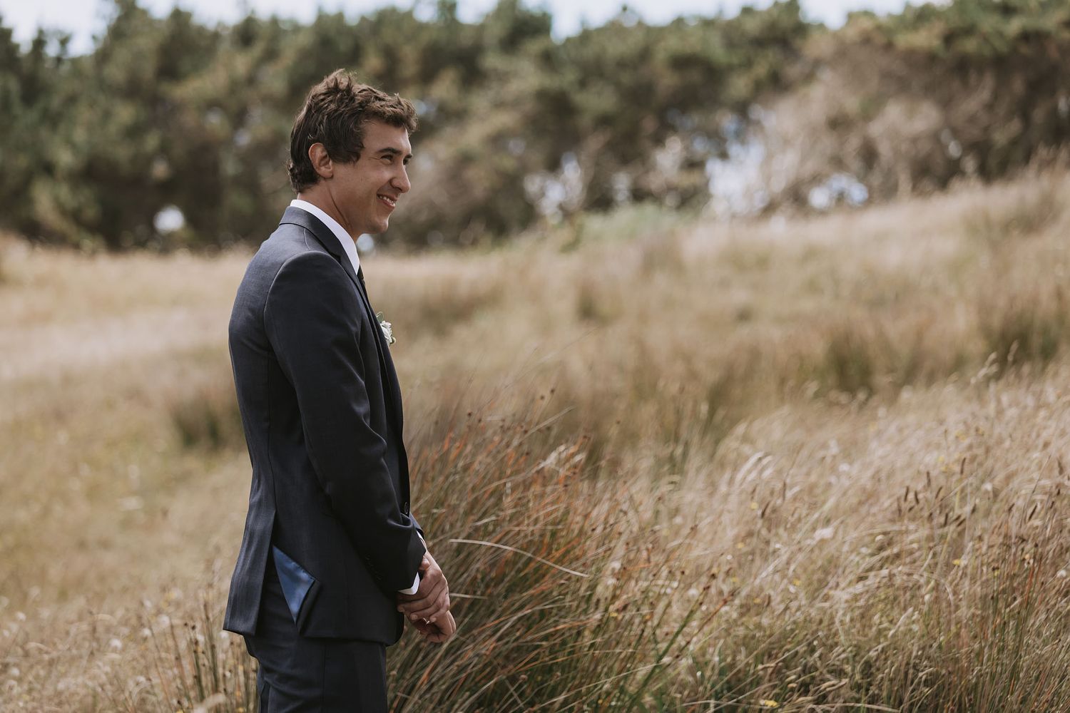 Person in a dark suit stands thoughtfully in a field of tall golden grass with trees in the background.
