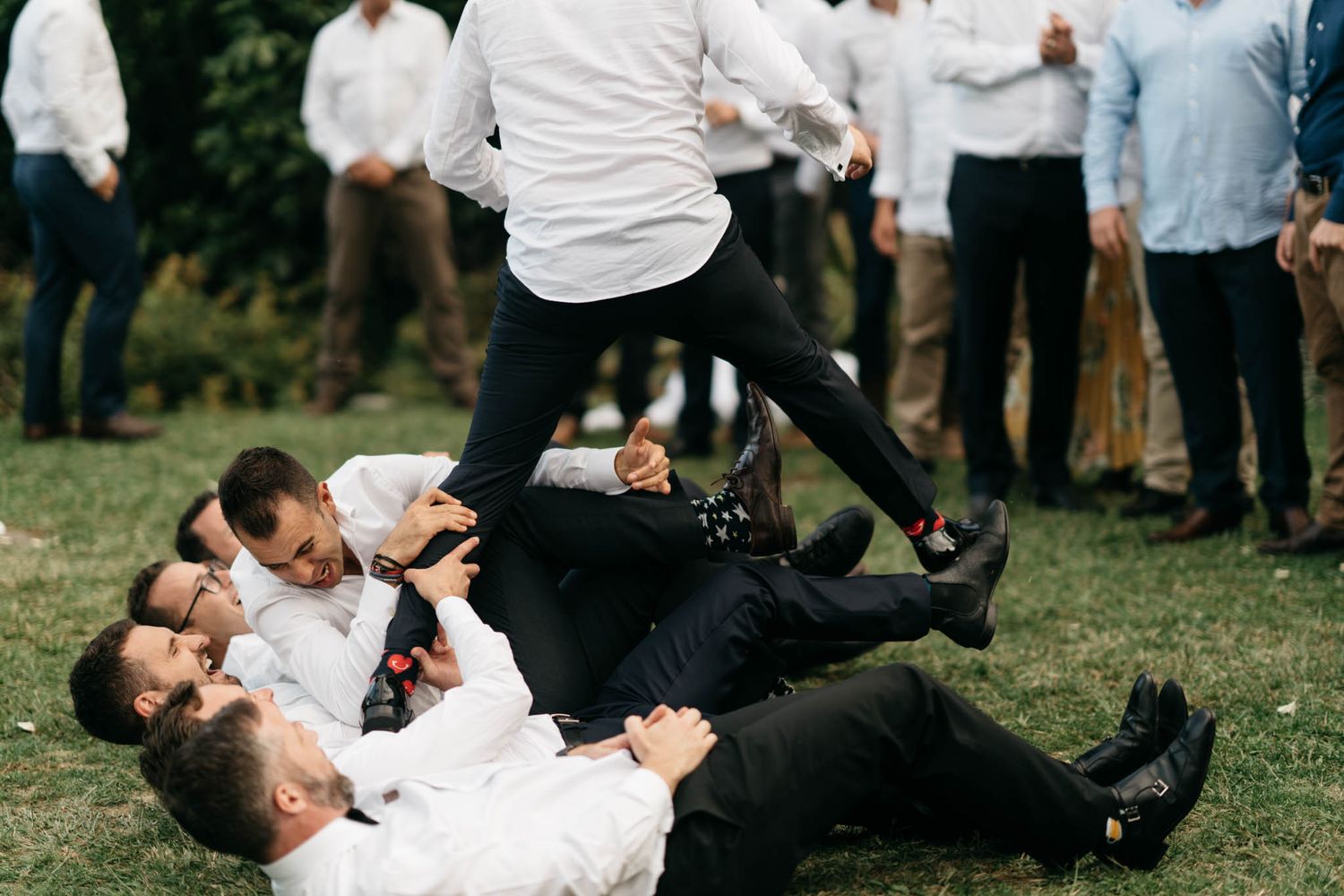 A group of people in white shirts and black suits playfully roll together on green grass during an outdoor gathering.