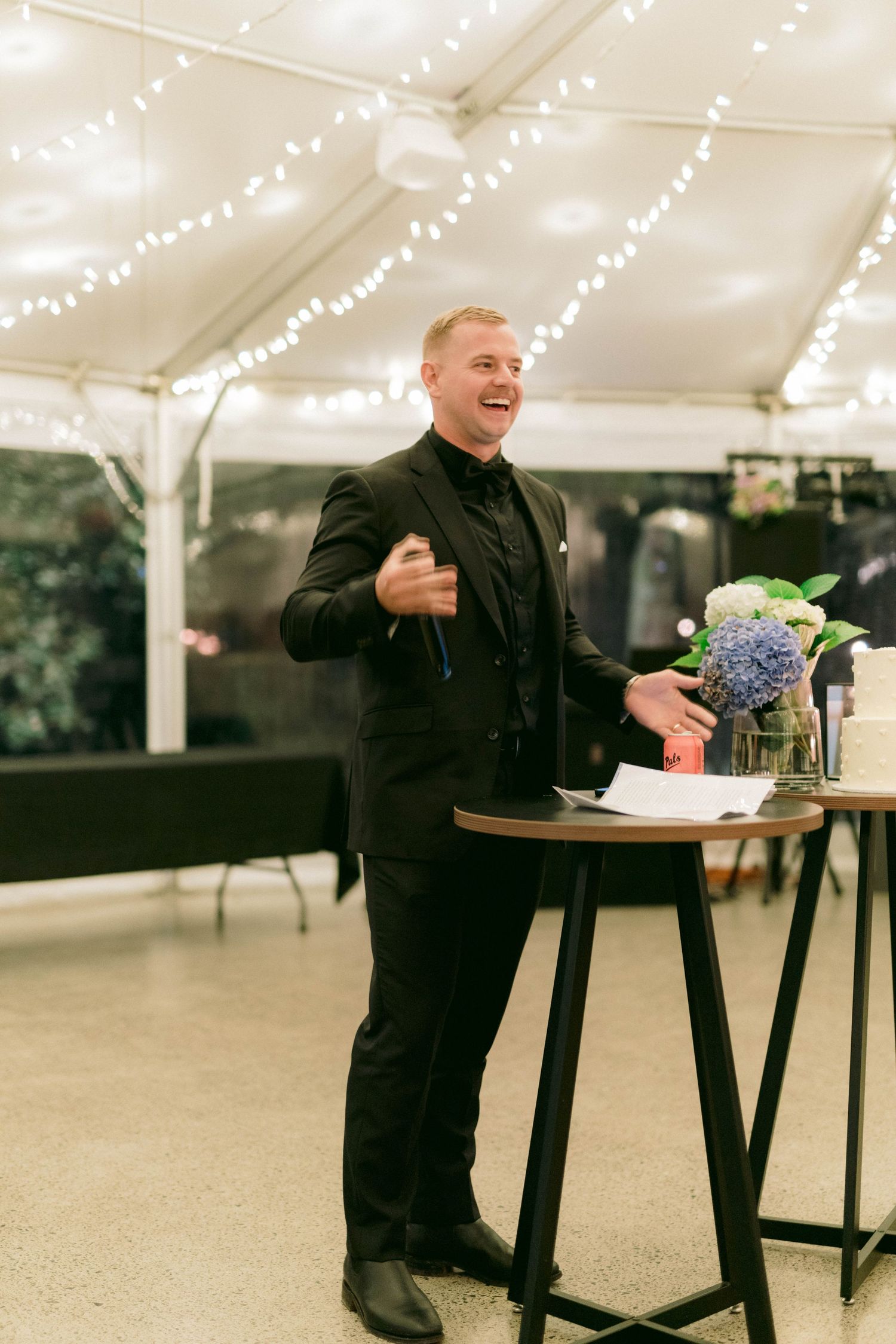A person in black attire delivers an animated speech at a cocktail table under a white tent with string lights.