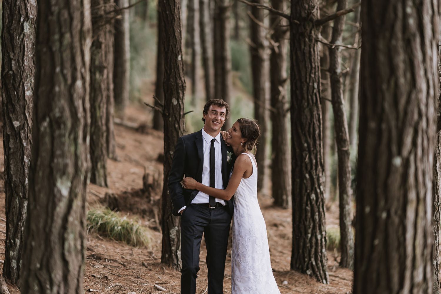 A couple in wedding attire stands among tall pine trees in a romantic forest setting.
