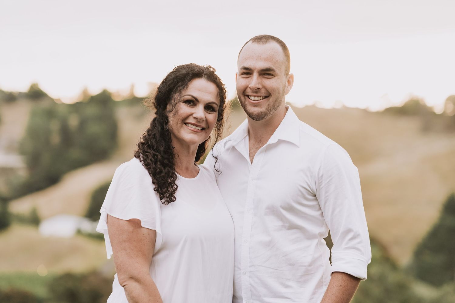 A couple in matching white shirts smiles while posing together outdoors during golden hour on a grassy hillside.