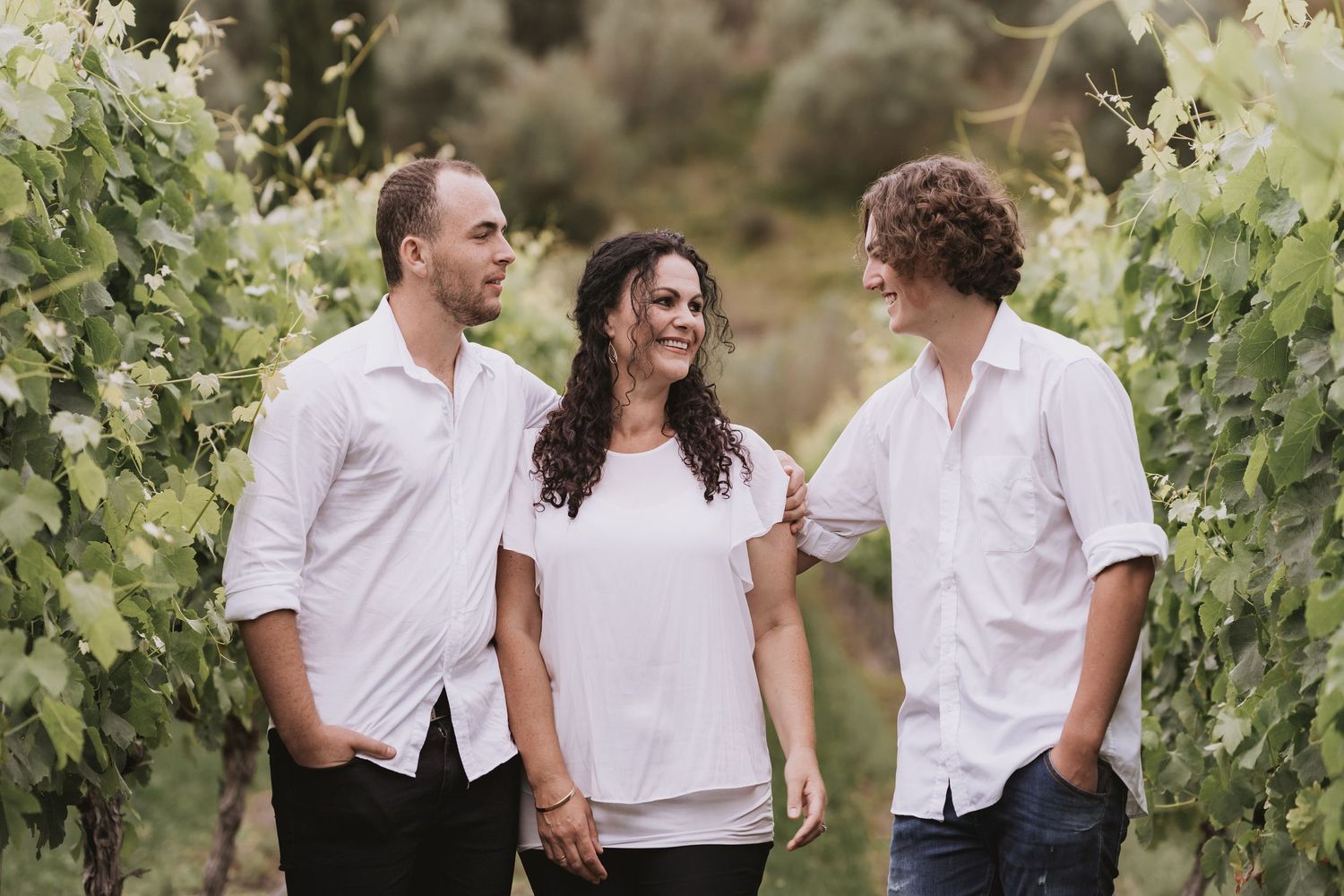 Three people in white shirts share a casual moment while standing among grape vines in a vineyard.