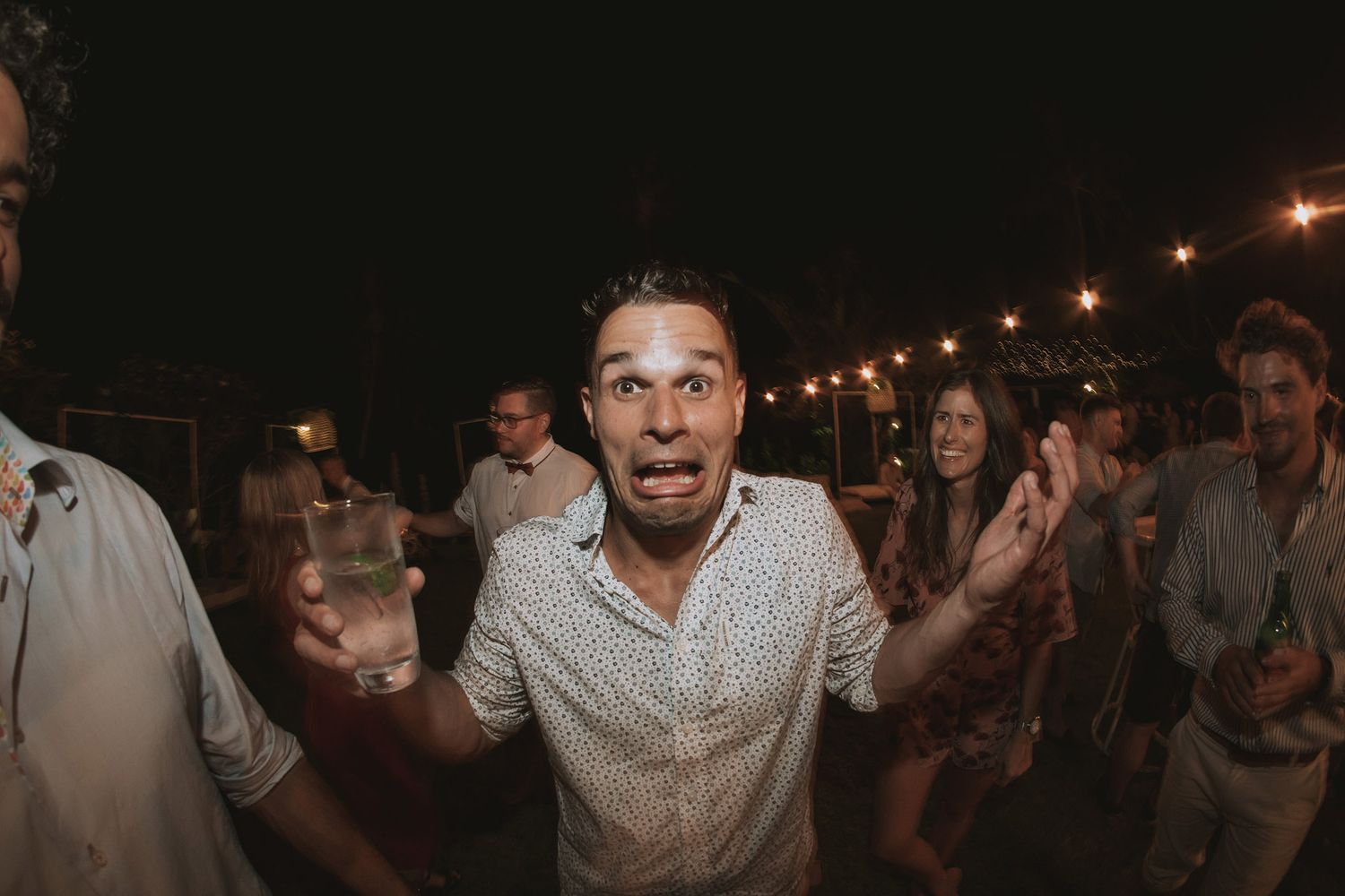 A person dances enthusiastically at a nighttime party under string lights, surrounded by other partygoers in the background.