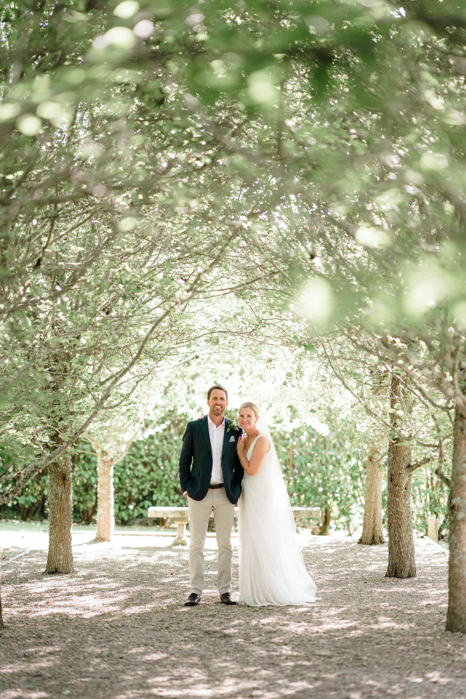 Newlyweds stand under tree-lined pathway in romantic outdoor garden setting.