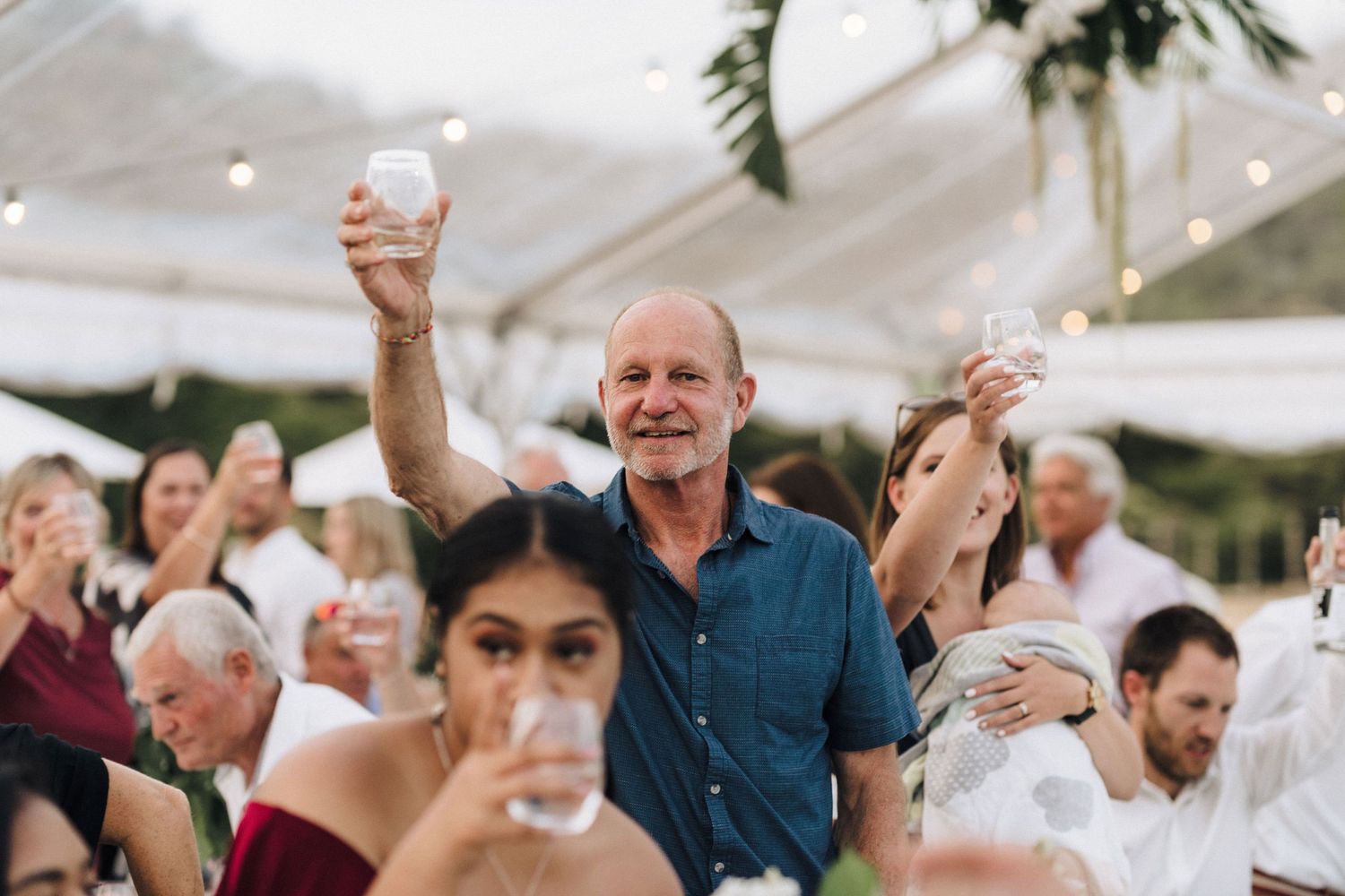 Wedding guests raising glasses in a toast under string-lit tent at dusk.