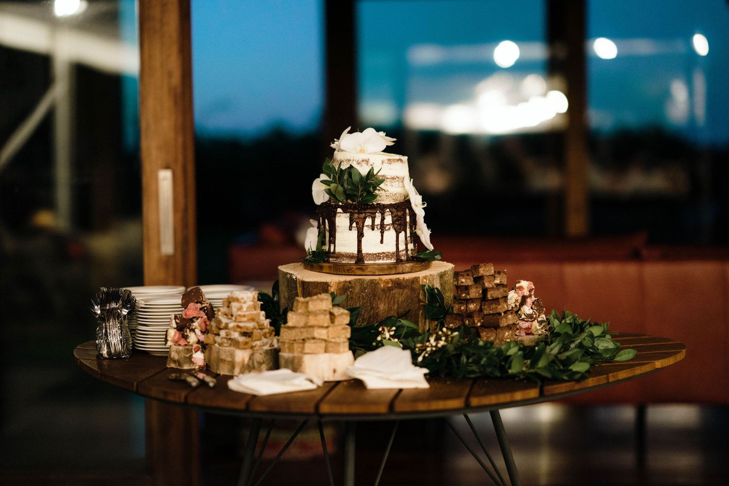 Elegant wedding dessert display with cake and greenery on wooden table at night.