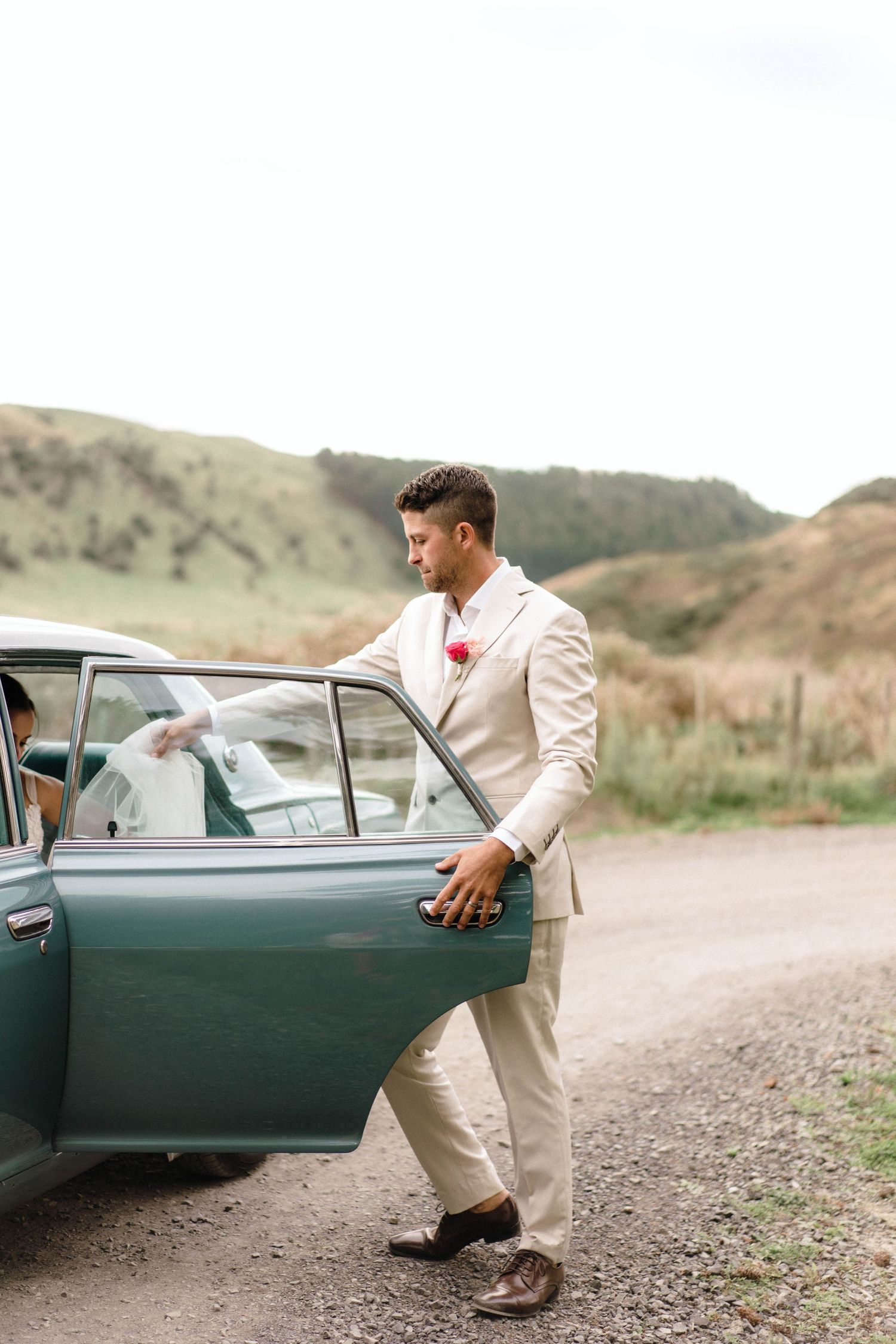 Wedding party interacting with vintage green car on dirt road.