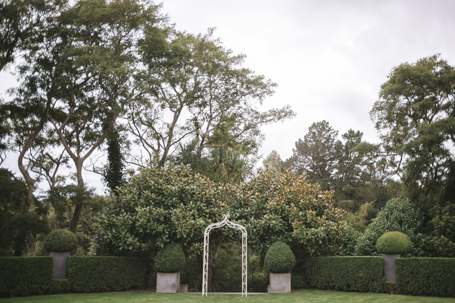 A serene garden path bordered by tall green hedges leads to an elegant white archway in a formal estate setting.