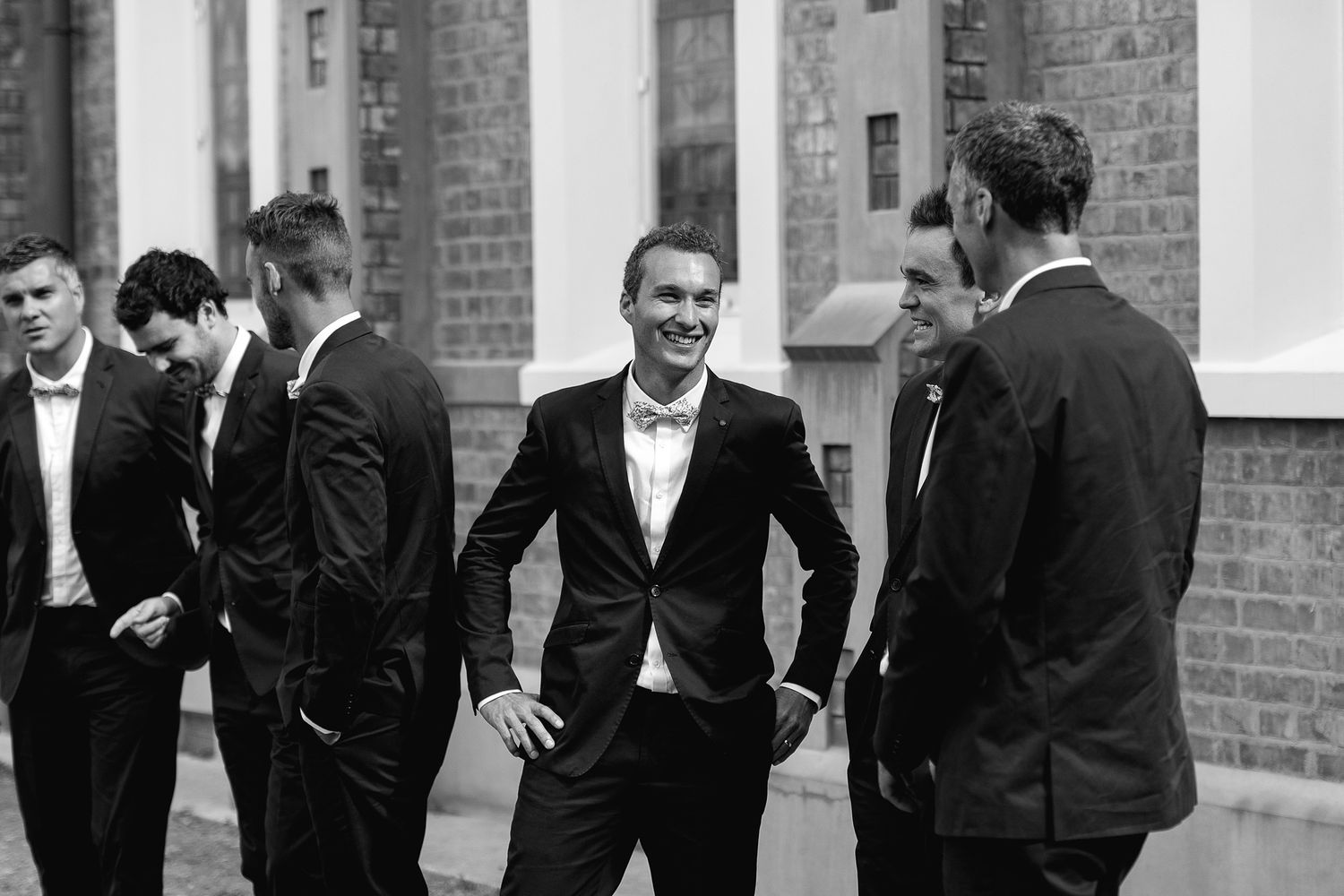 A black and white photograph shows a line of formally dressed men in suits standing against a brick wall.