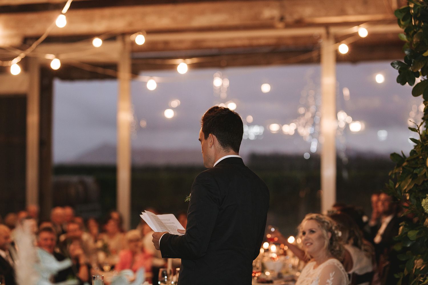 Best man delivers wedding speech at evening reception under string lights at outdoor covered venue.
