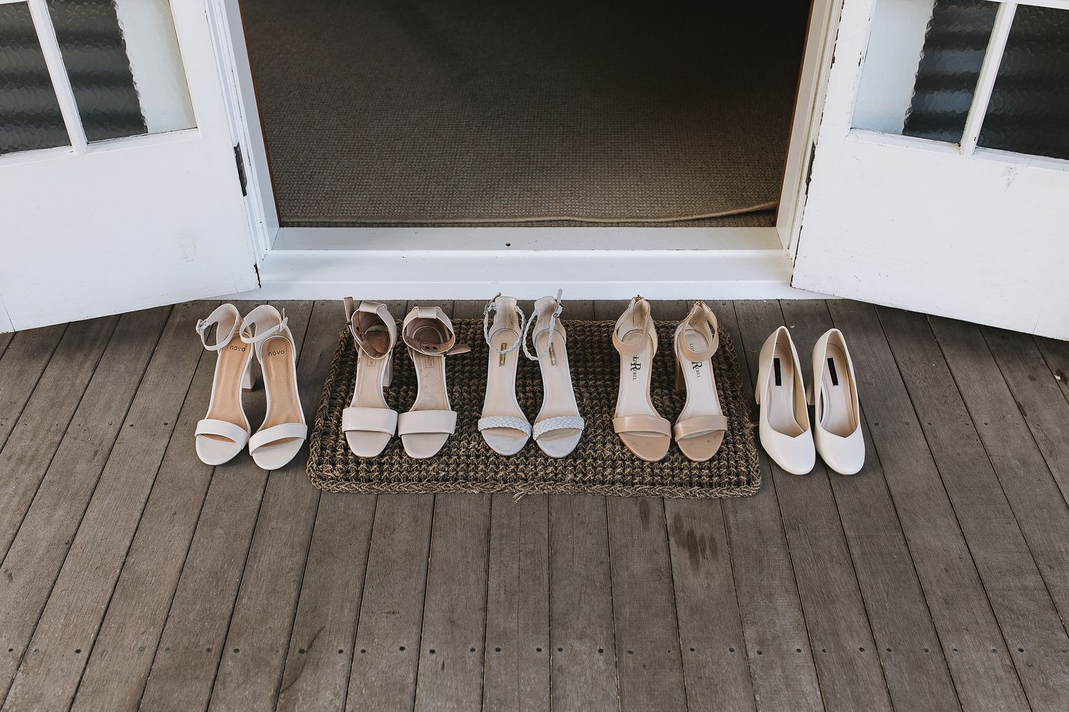 A row of light colored formal women's heels neatly arranged on a wooden porch floor against white French doors.
