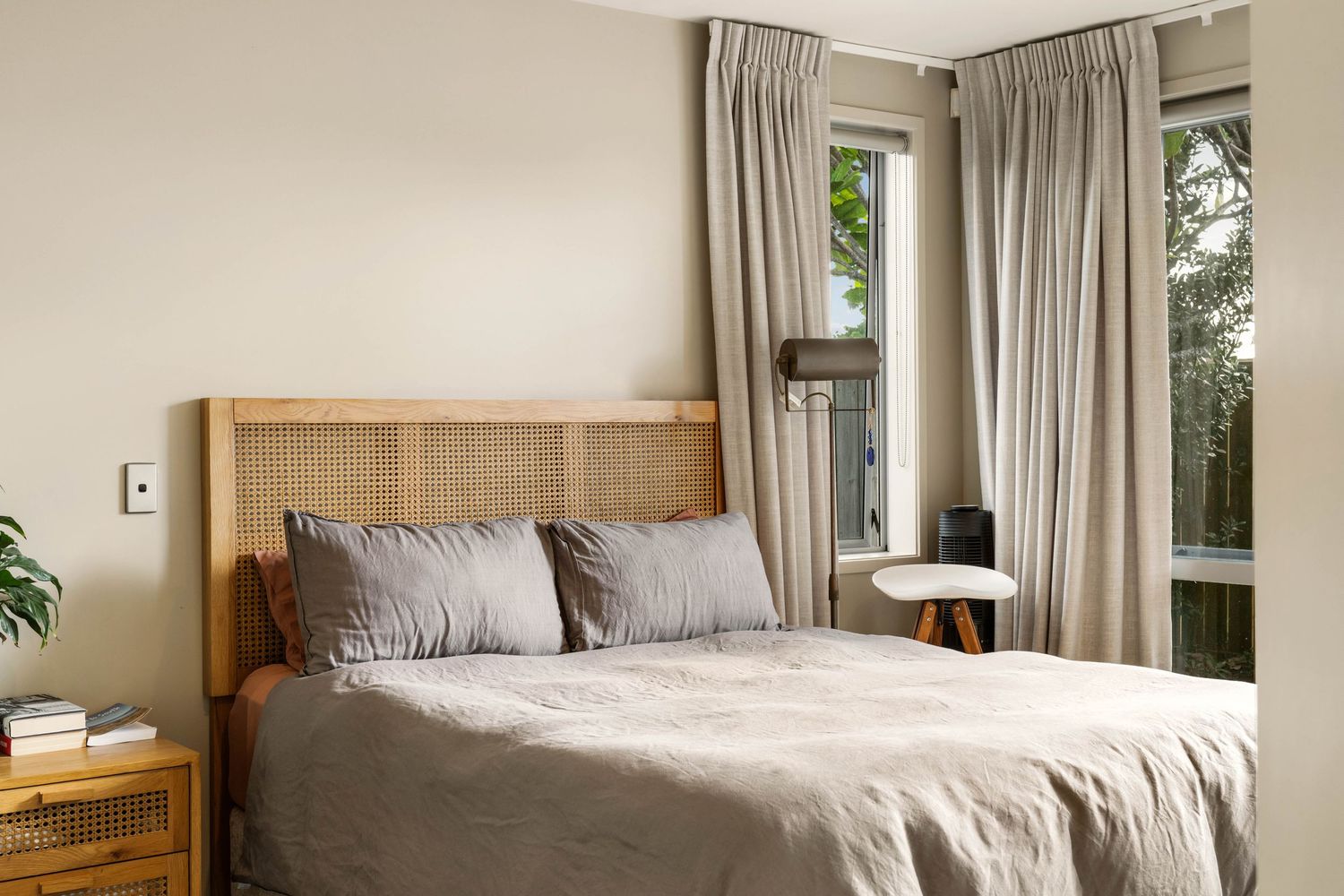 A minimalist modern bedroom with light beige curtains, wooden bed frame, and natural sunlight streaming through windows.