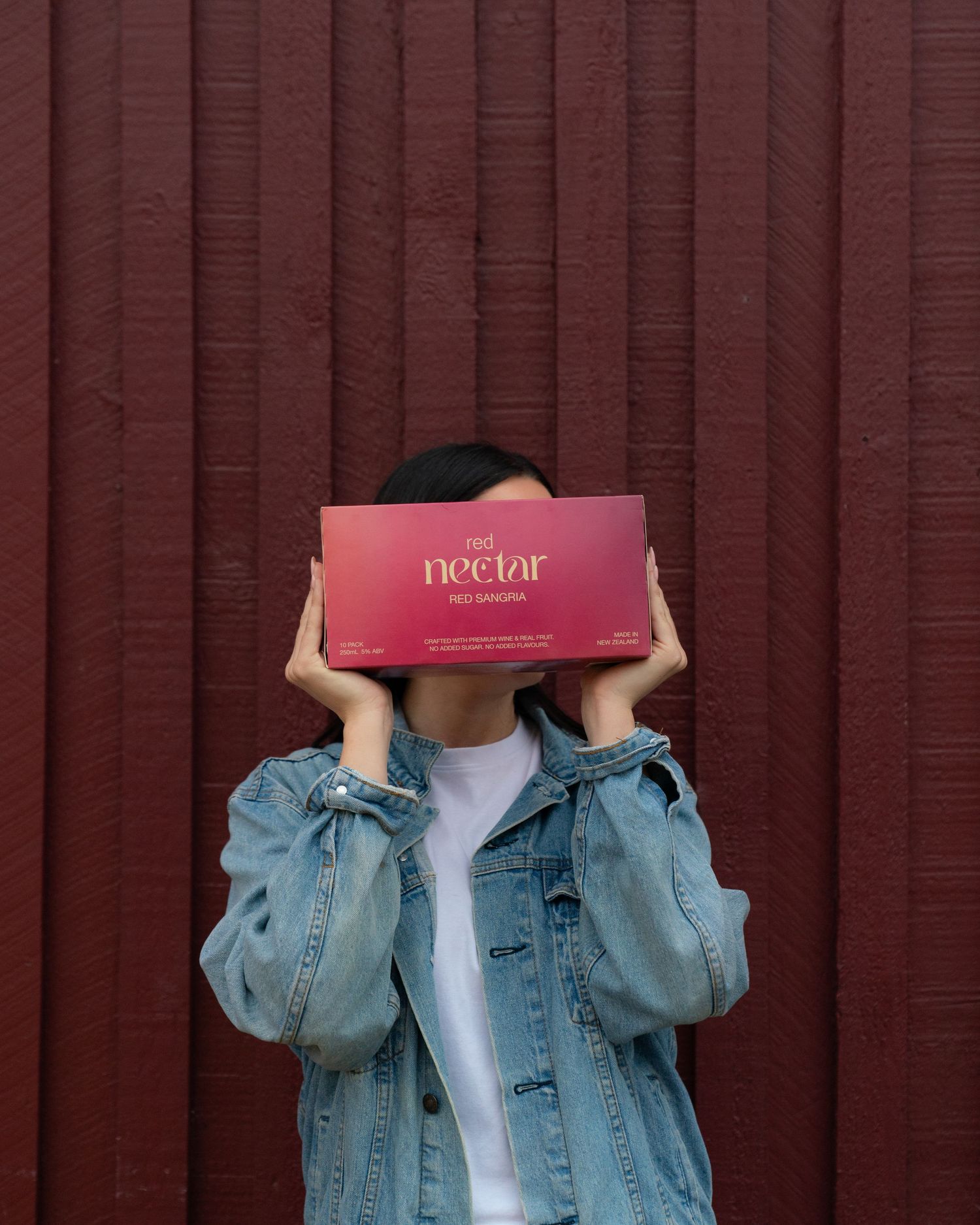 A person holding a pink Nectar can in front of their face while wearing a denim jacket against a red wooden wall.