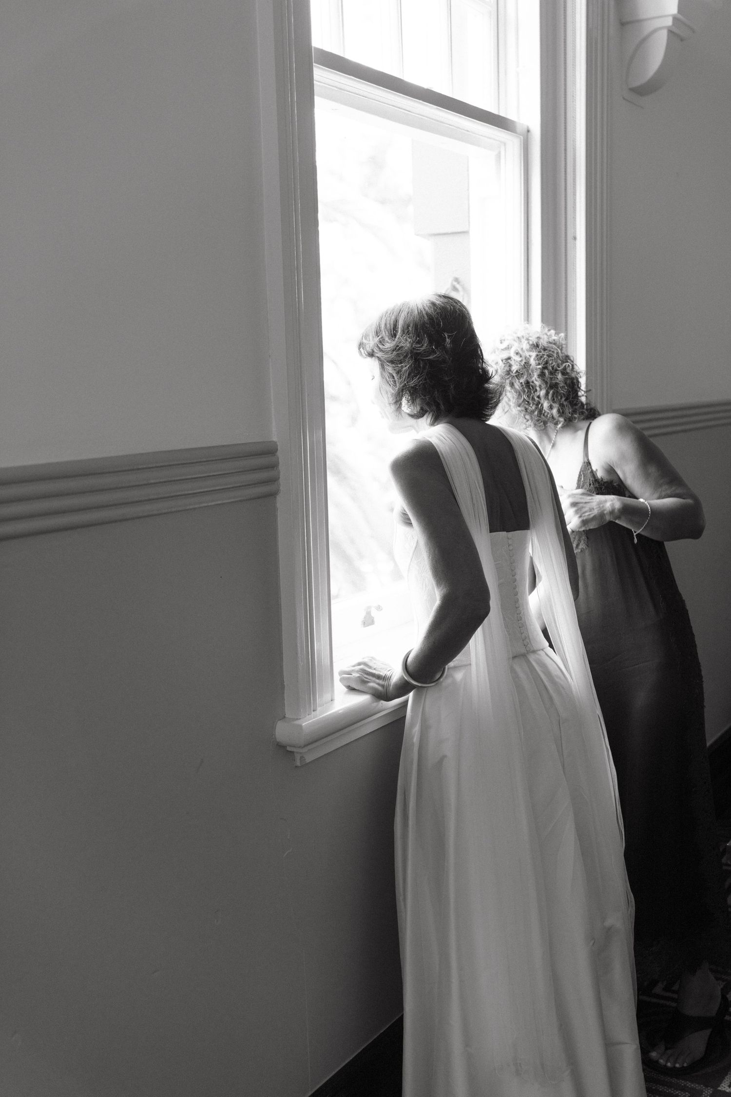 A black and white photo of two people in formal attire looking out of a tall window.