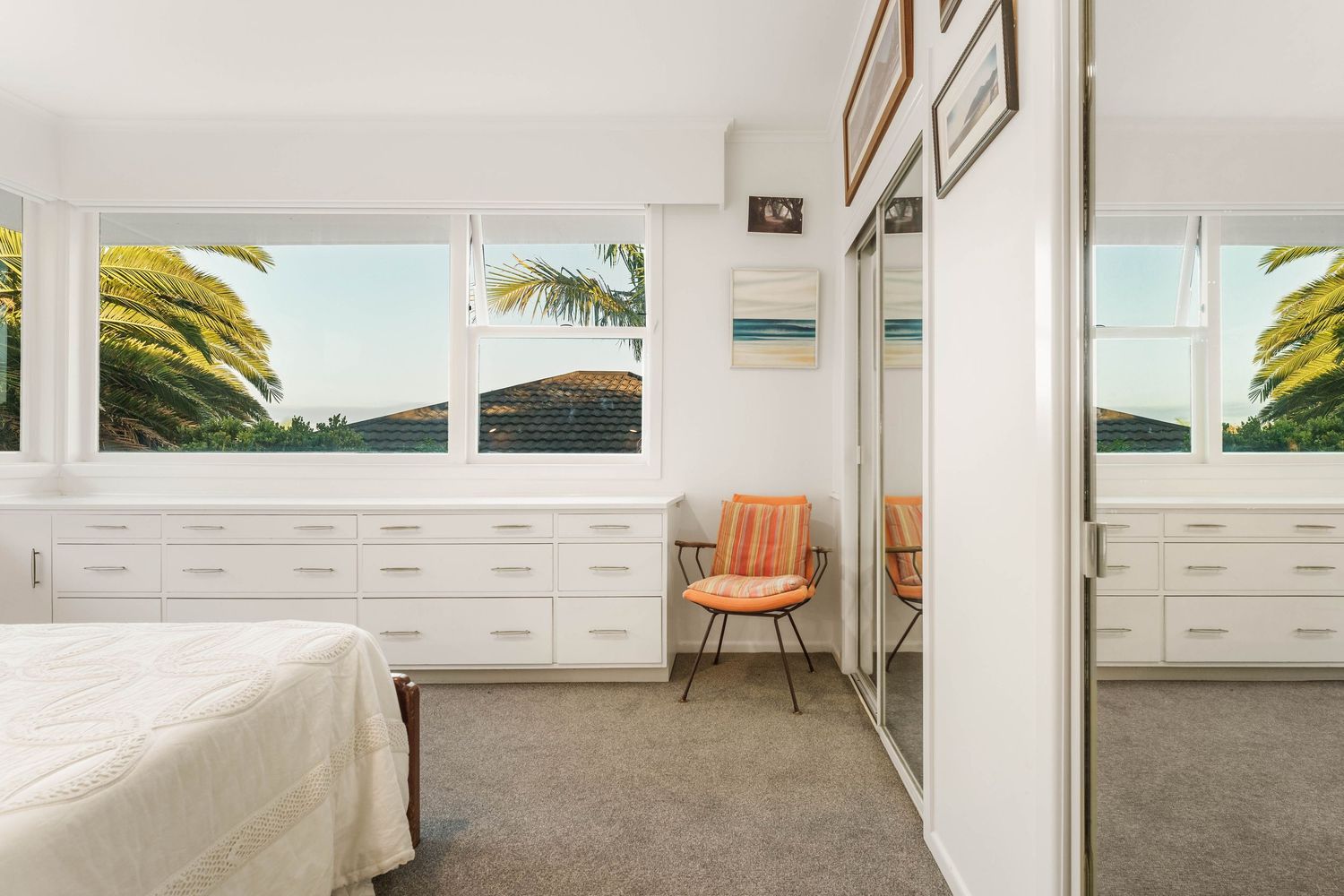 Bright coastal bedroom with white furniture, orange accent chair, and large windows overlooking palm trees and ocean views.