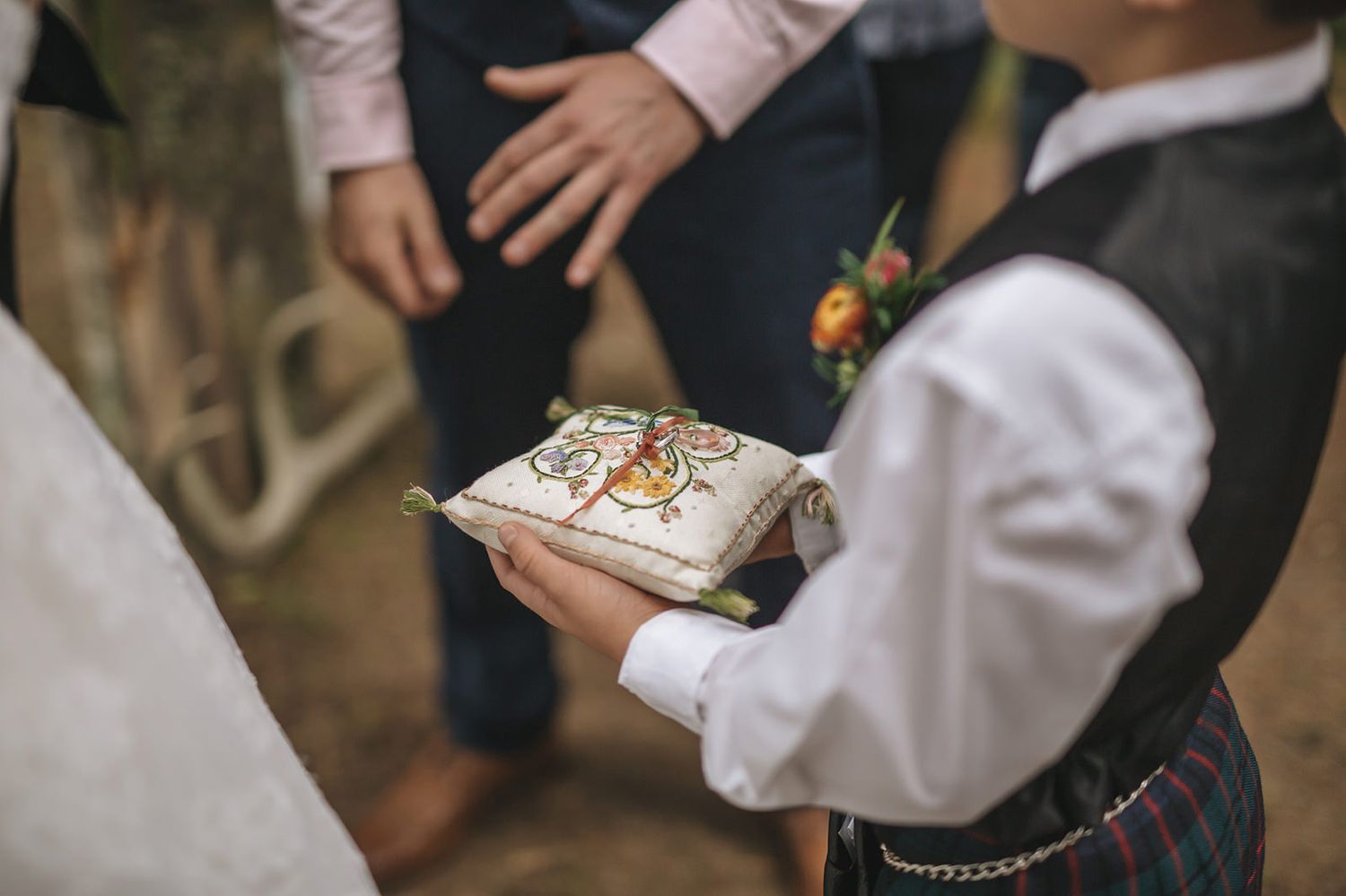 A ring bearer in a white dress shirt holds a decorative pillow with wedding rings during a ceremony.