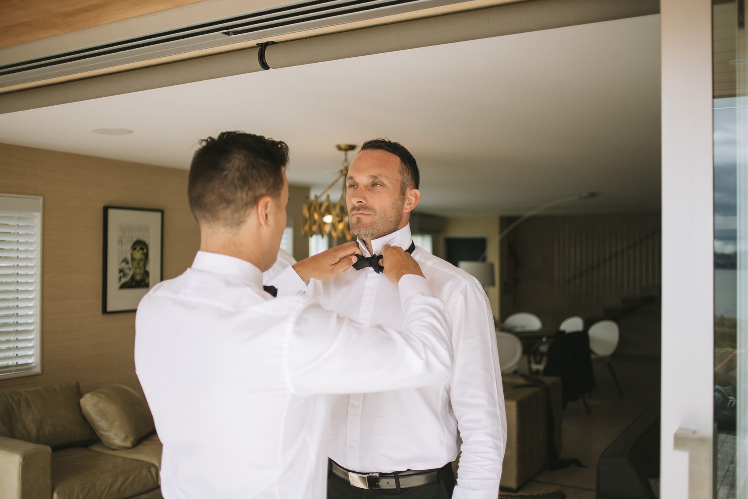 A groom gets ready for his wedding day with groomsmen helping adjust his tie and boutonniere in a sunlit room.