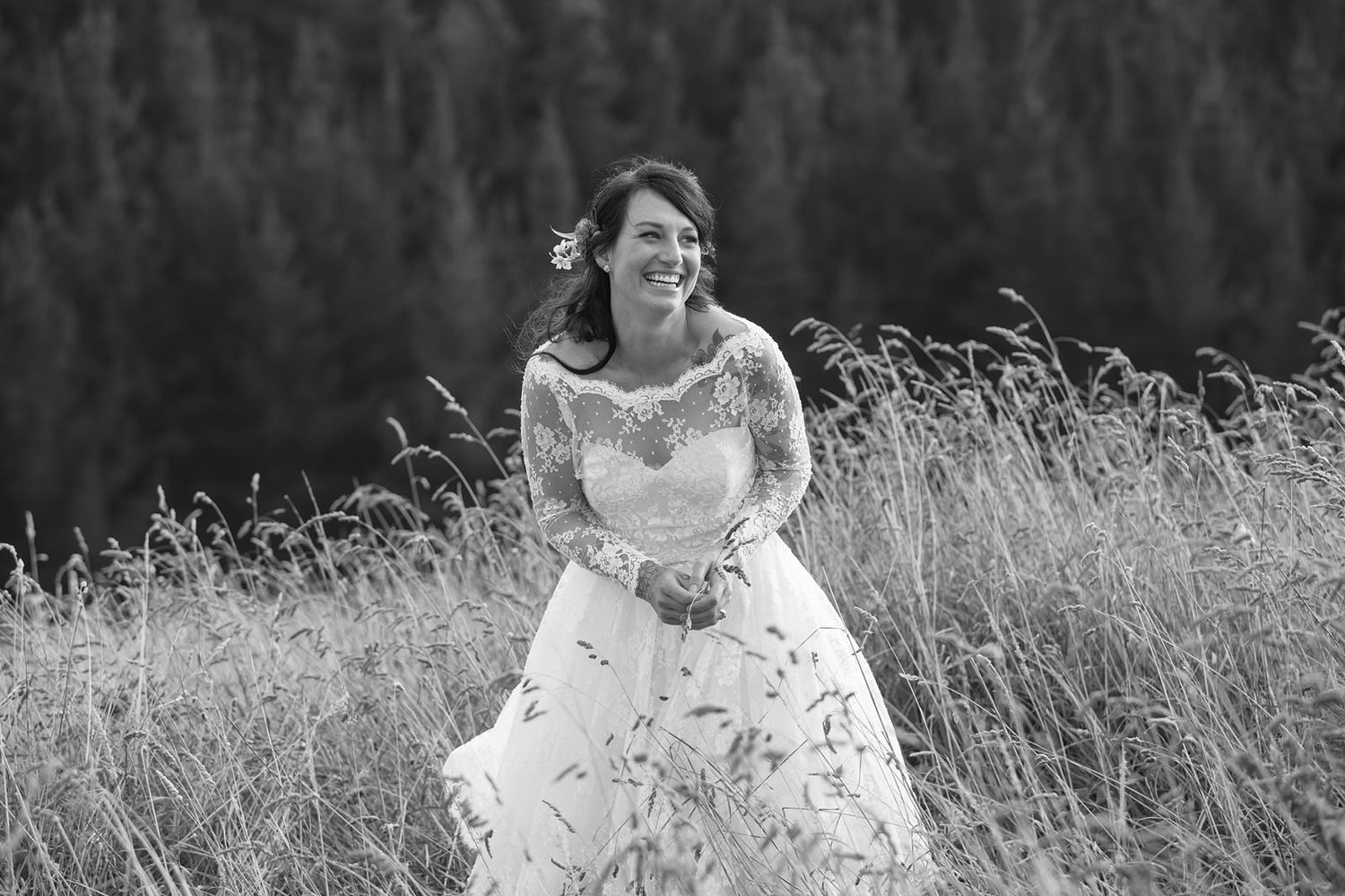 A person in a white lace wedding dress walks through a scenic field of tall grass and wildflowers.