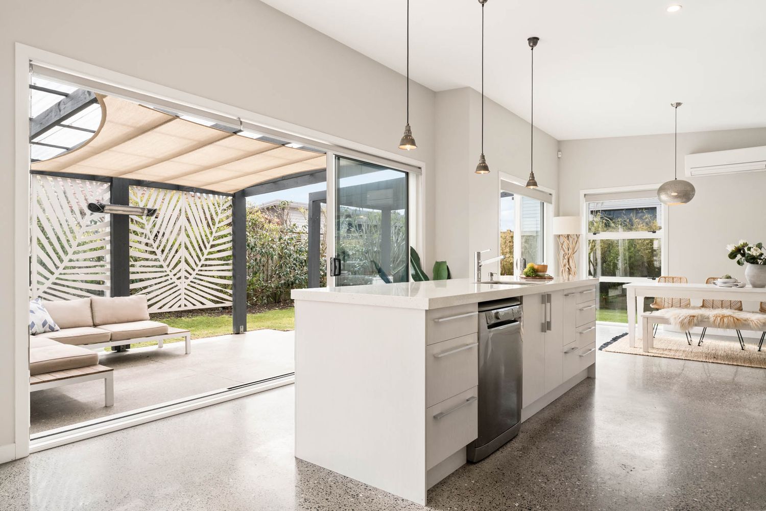 A sleek modern kitchen with minimalist white cabinets features pendant lighting and large windows overlooking an outdoor area.