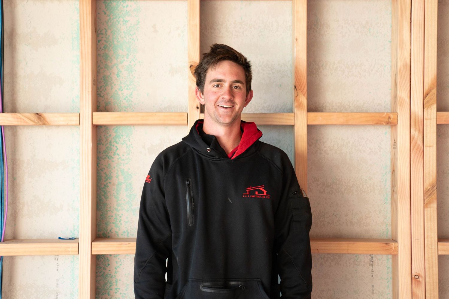Someone wearing a black hoodie stands against a wooden panel wall in an unfinished room.
