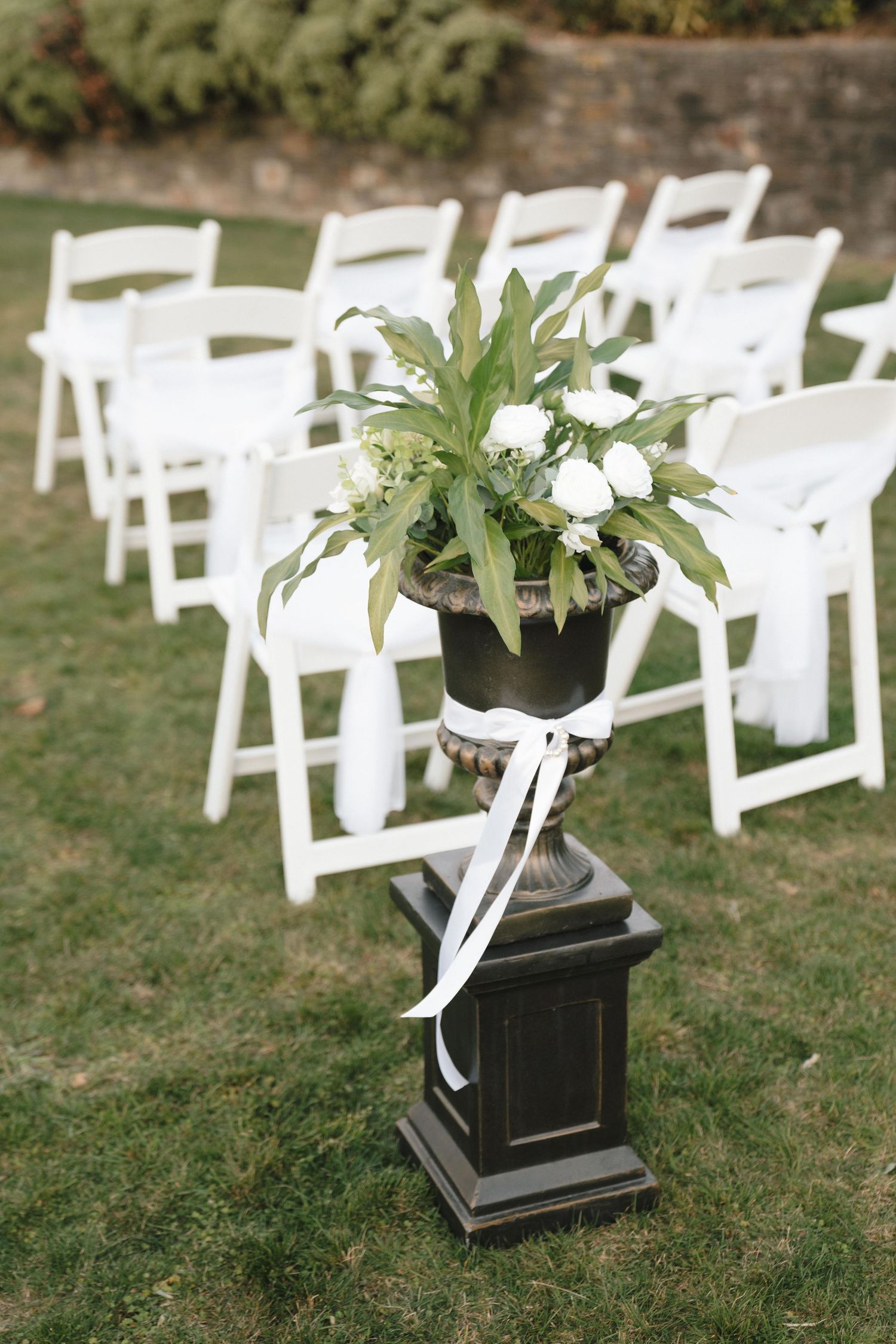 White garden chairs arranged in rows with a floral arrangement on a black pedestal for an outdoor ceremony.