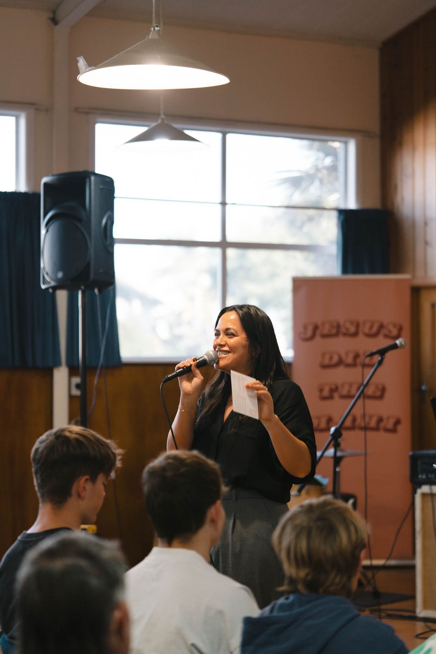 A speaker holds a microphone while addressing an audience in a room with wooden walls and audio equipment.