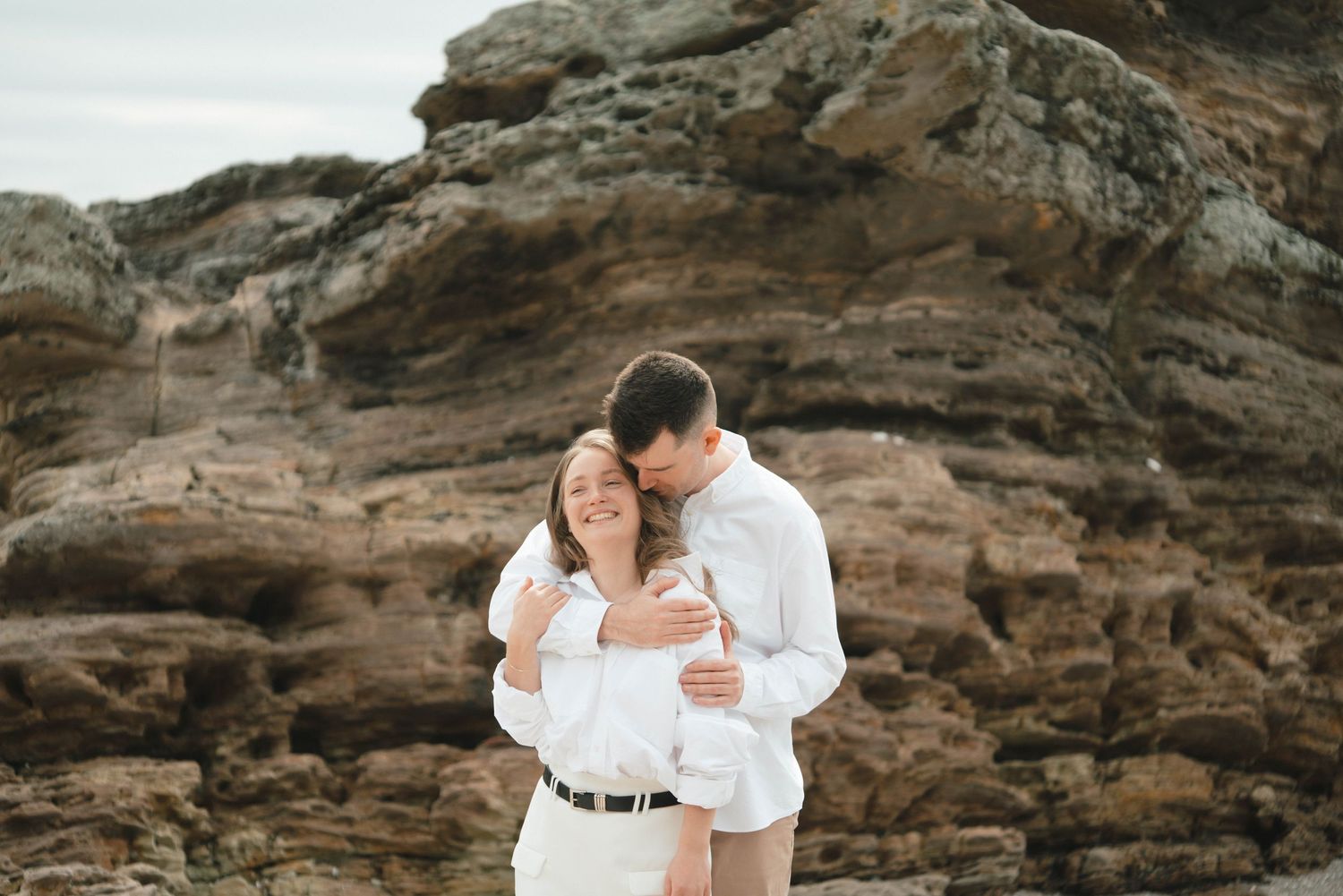A romantic couple in white outfits embraces against rugged coastal rocks during a photoshoot at the beach.
