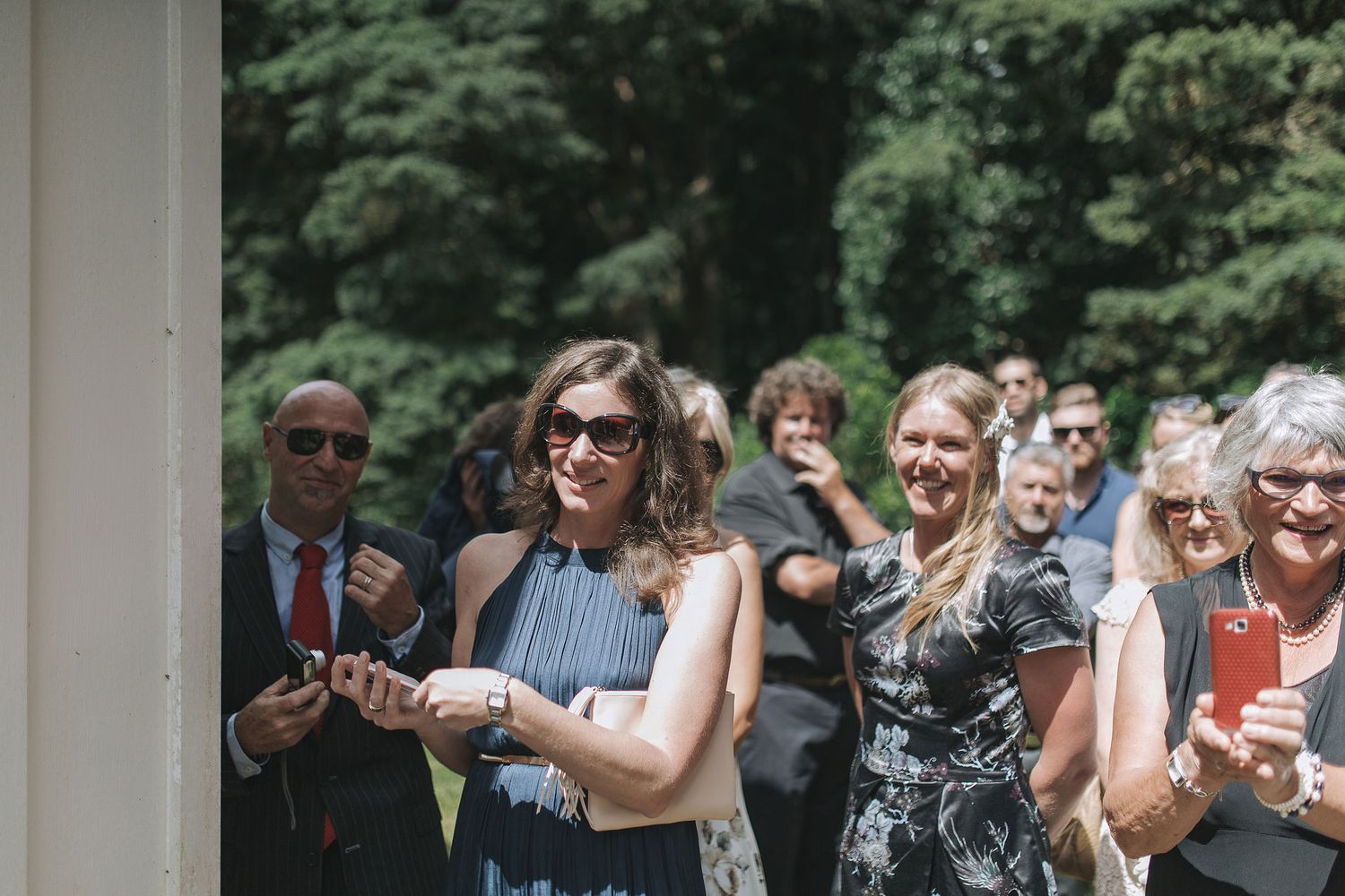 Wedding guests in formal attire mingle outdoors under a white columned structure during a daytime celebration.