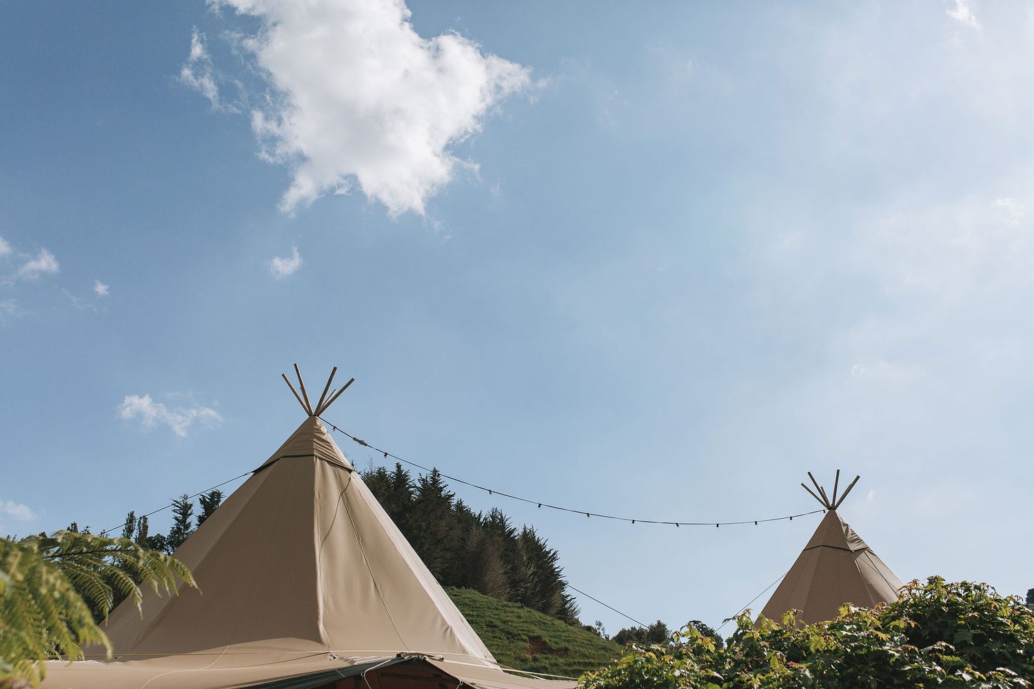 Two tan tipi tents with pointy tops set up outdoors against a blue summer sky with white fluffy clouds.