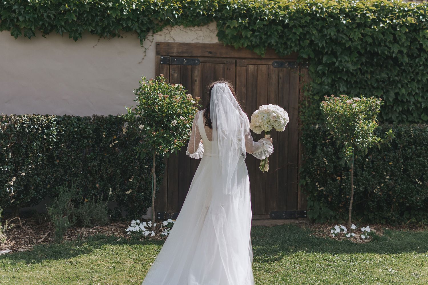 A bride in a white wedding gown stands in a lush garden setting with a floral bouquet against greenery.