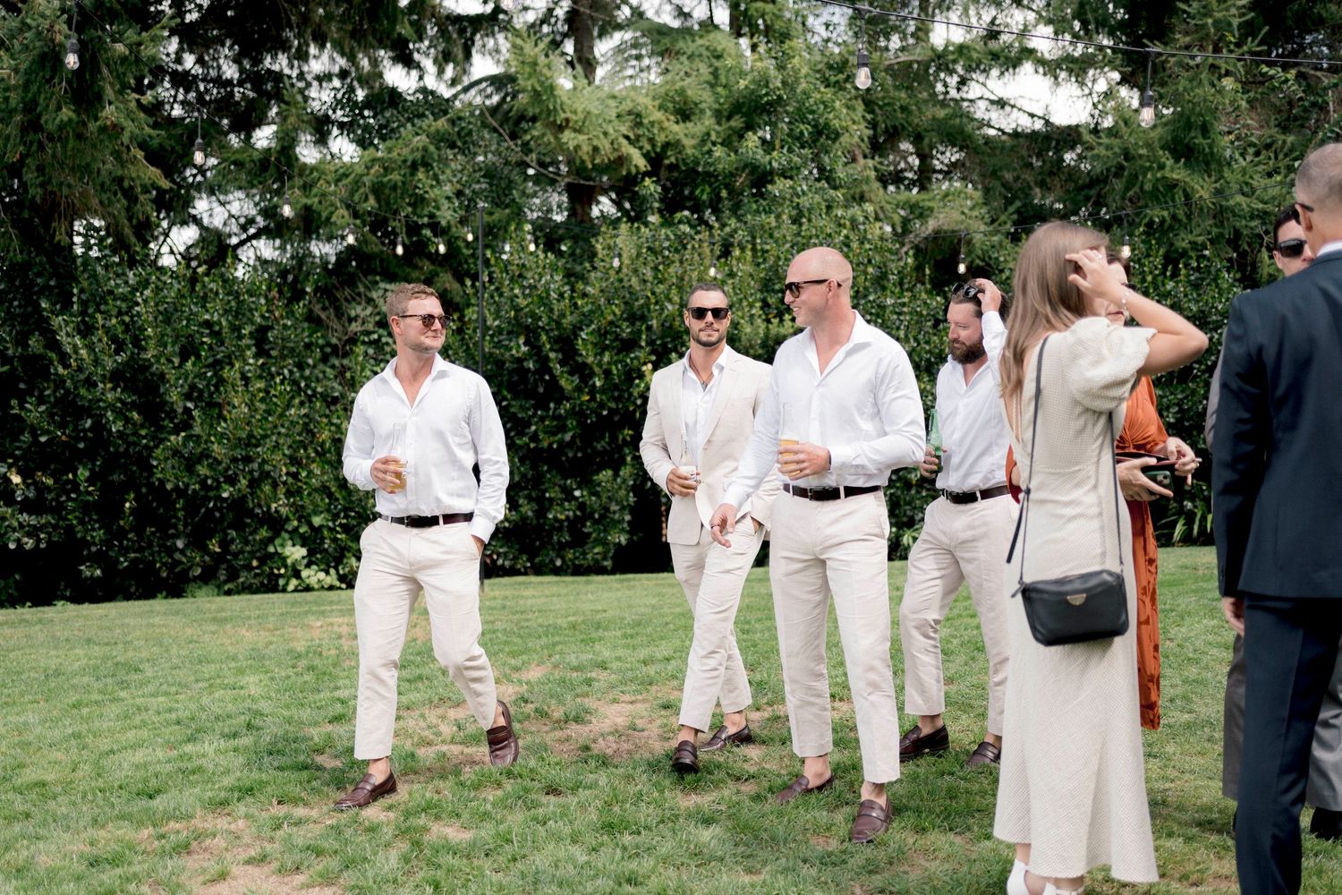 Wedding guests in light colored attire gather on a grassy lawn overlooking water.