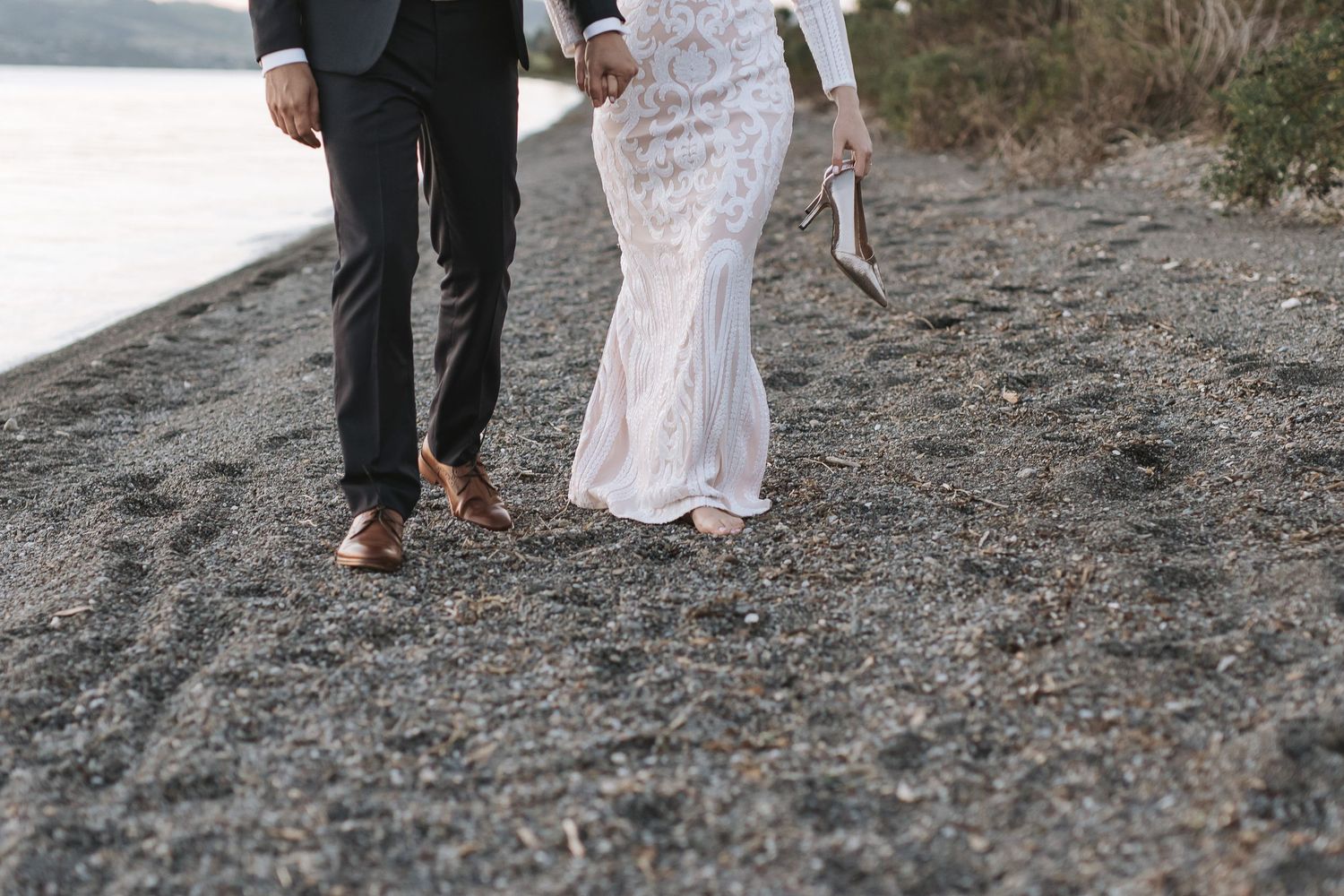 A couple walks along a rocky beach shoreline with the person wearing a flowing white lace wedding dress.