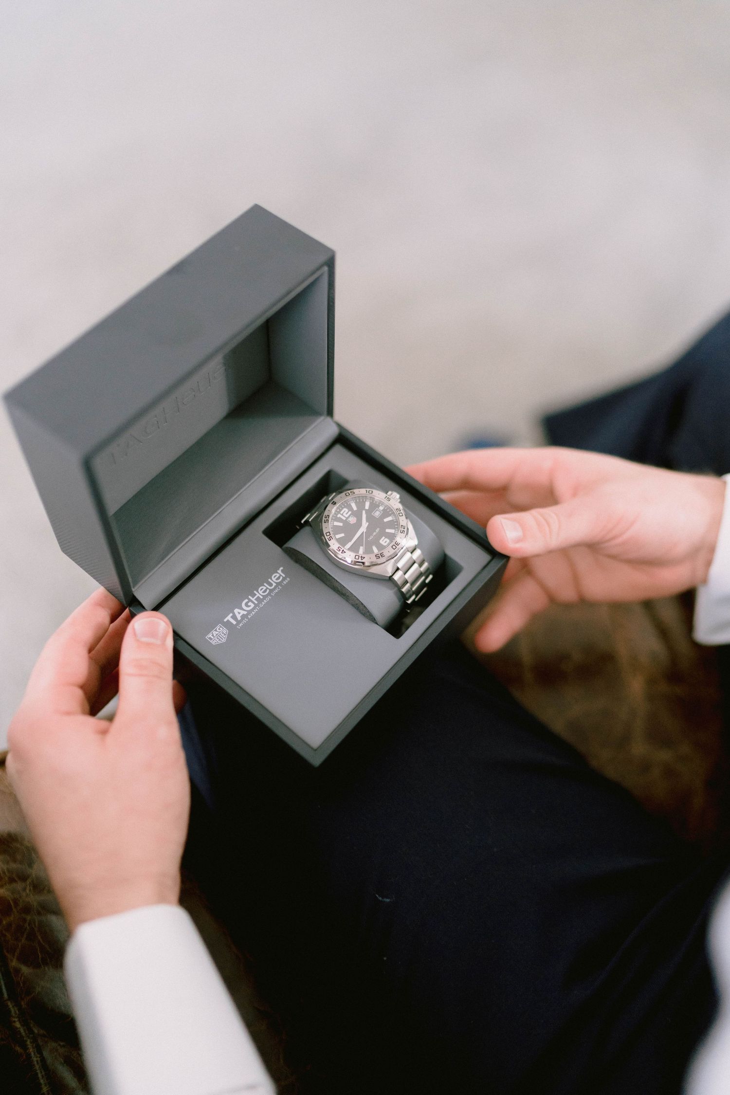 Sequence of hands opening a sleek black Tag Heuer watch box to reveal a silver luxury timepiece inside.