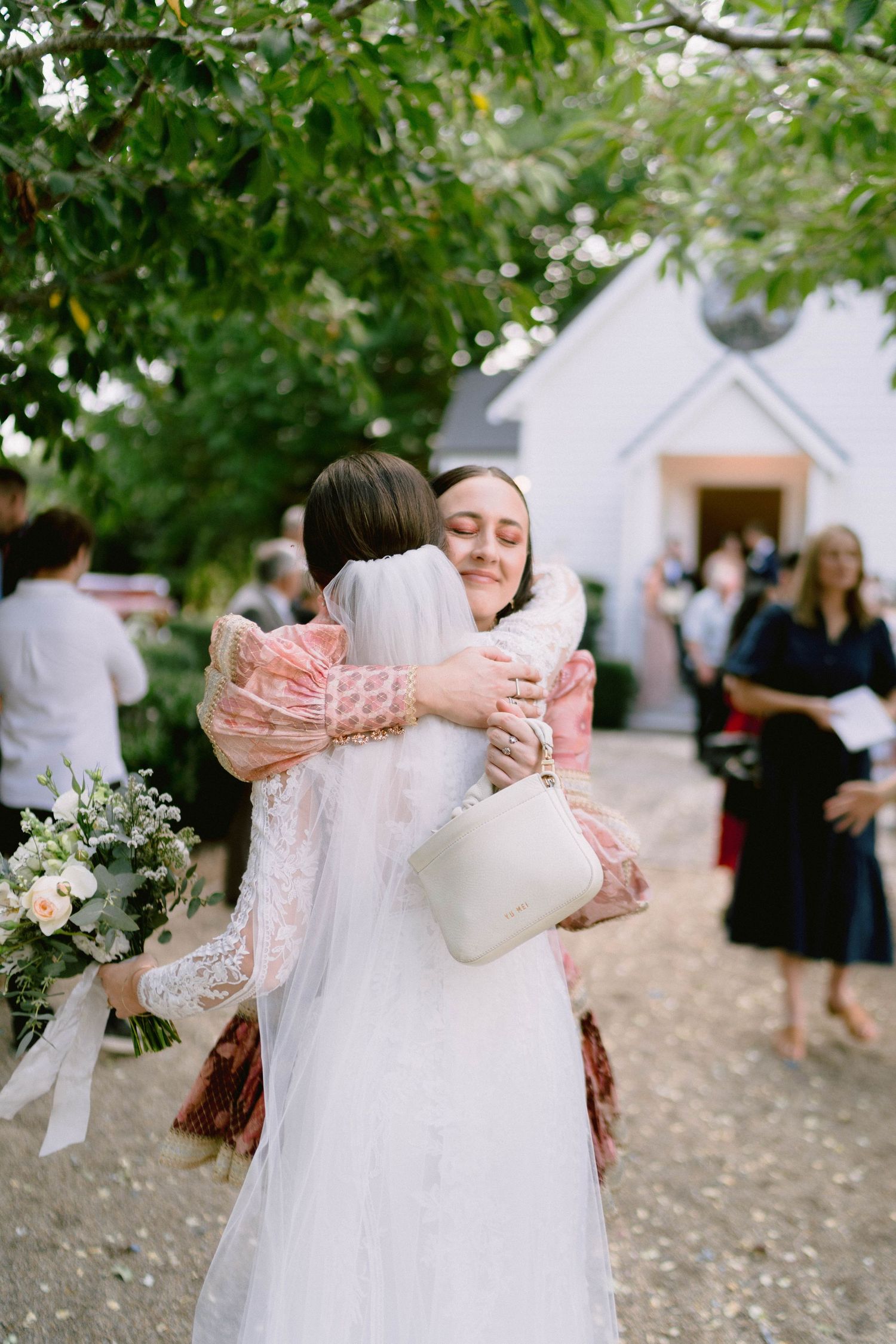 A warm embrace between wedding guests outside a charming white chapel surrounded by greenery.