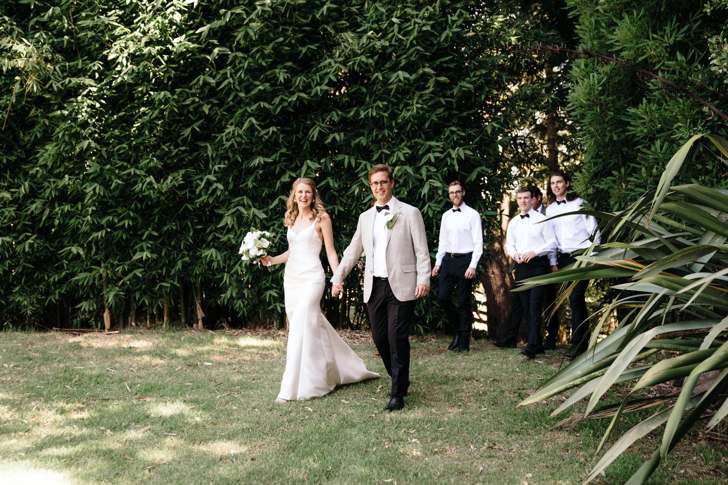 Wedding party stands together on grassy lawn surrounded by lush green trees and plants for outdoor garden photos.